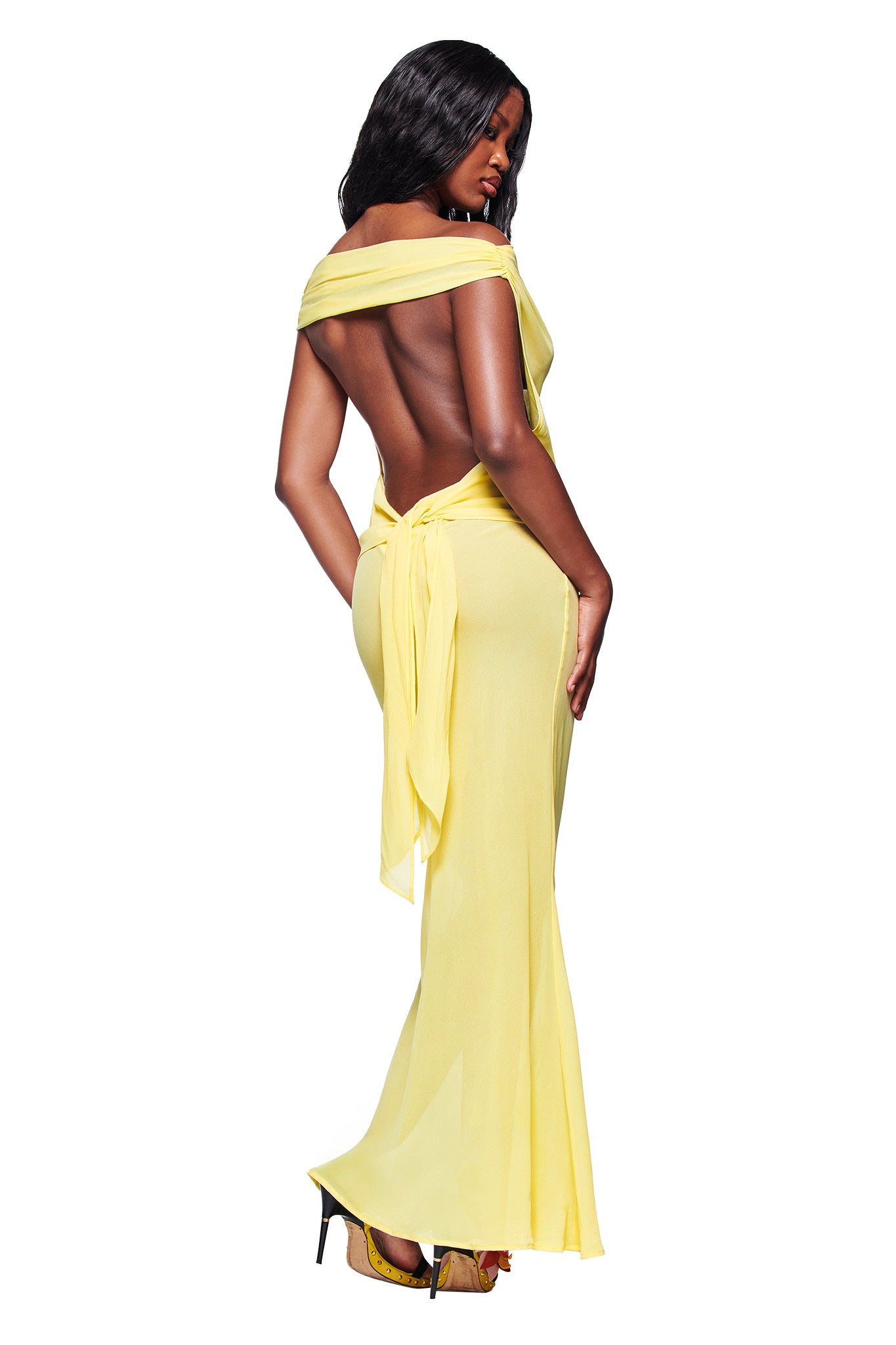 A woman poses with her back to the camera in the I.AM.GIA KAHLO MAXI DRESS - YELLOW, a lightweight georgette off-the-shoulder backless maxi dress, paired with high heels against a white background.