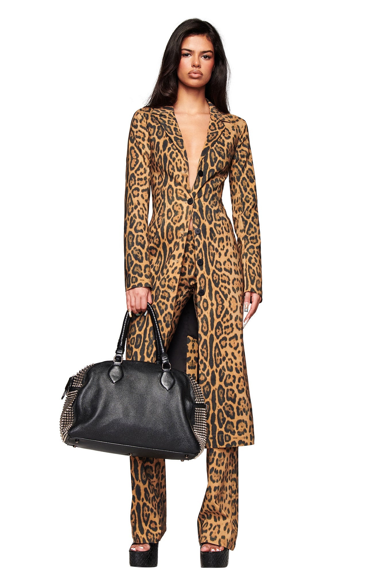 A woman faces forward in a longline I.AM.GIA UMA COAT - PANTHERA with matching leopard print pants, holding a large black handbag and wearing black platform shoes against a white background.