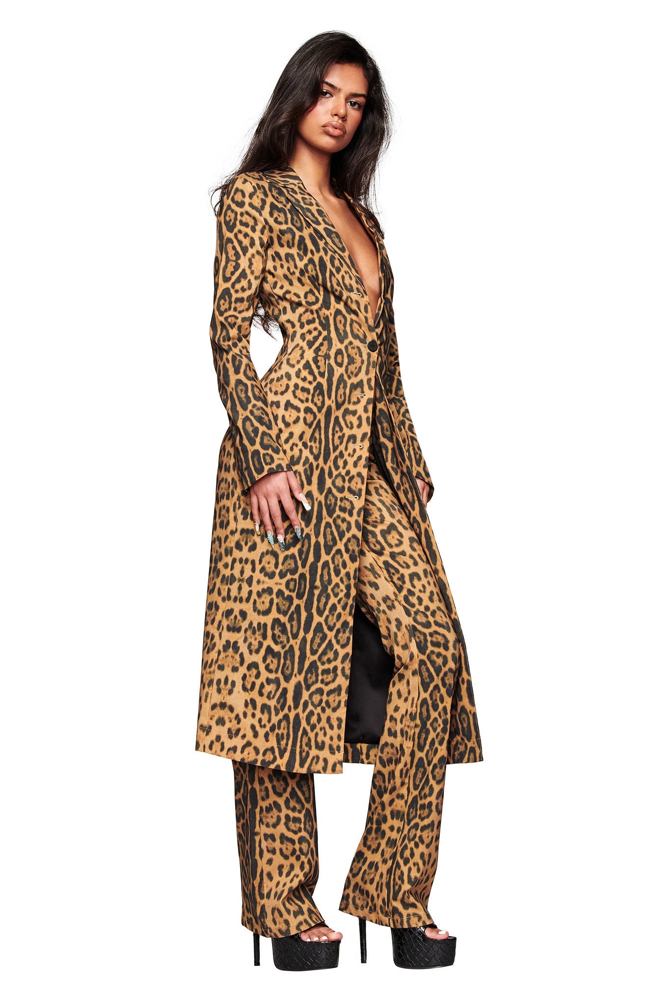 A woman models the I.AM.GIA UMA COAT - PANTHERA, a longline tailored animal print coat paired with matching wide-leg pants and platform heels. The bold leopard pattern and deep V-neck create a striking look against a white background.
