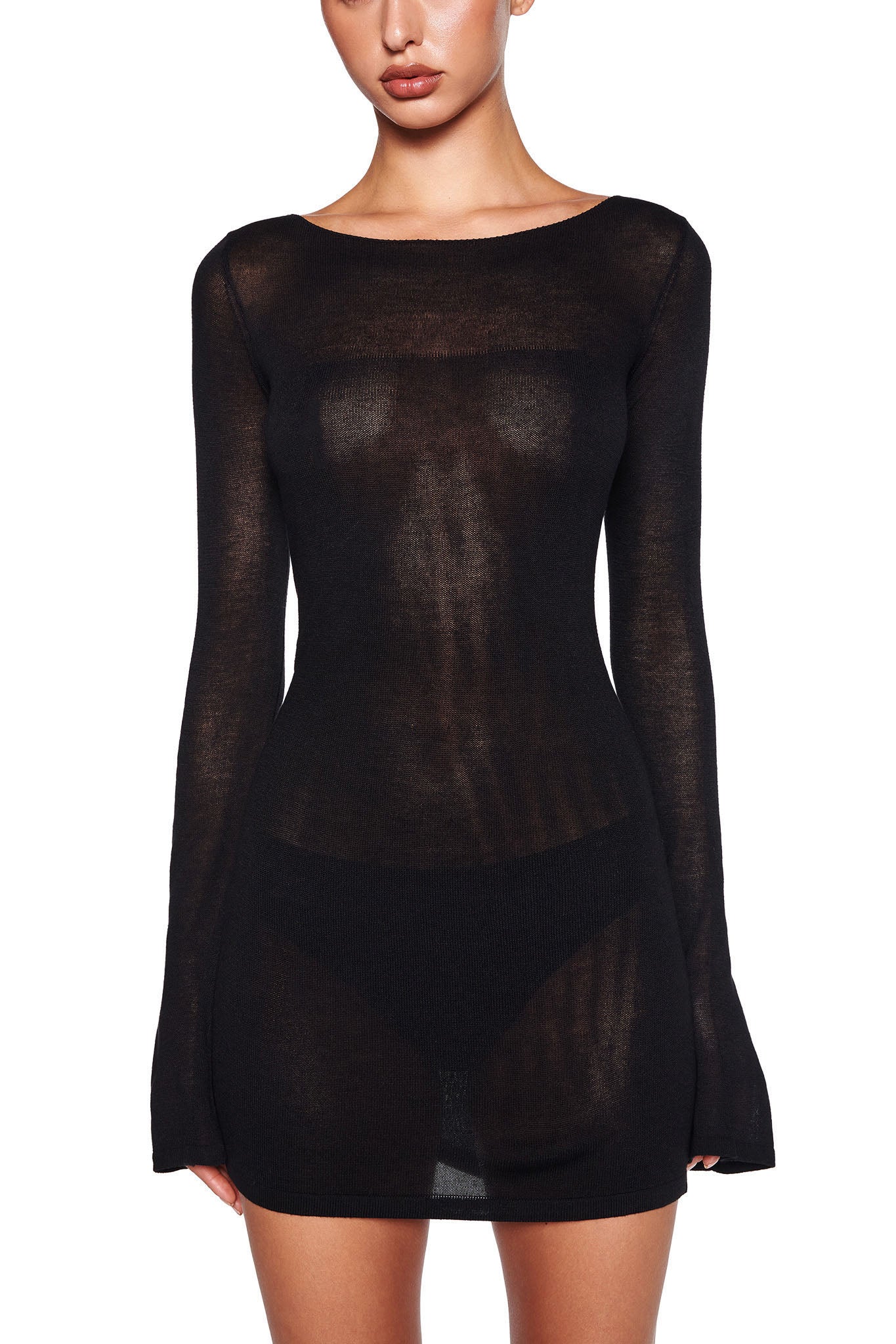 A woman is wearing the VIOLET KNIT DRESS - BLACK by I.AM.GIA. This sheer, long-sleeved dress is semi-transparent and form-fitting, featuring a modest boat neckline and an A-line silhouette. Against the white background, the black dress stands out prominently as the center of attention while her face remains partially hidden.