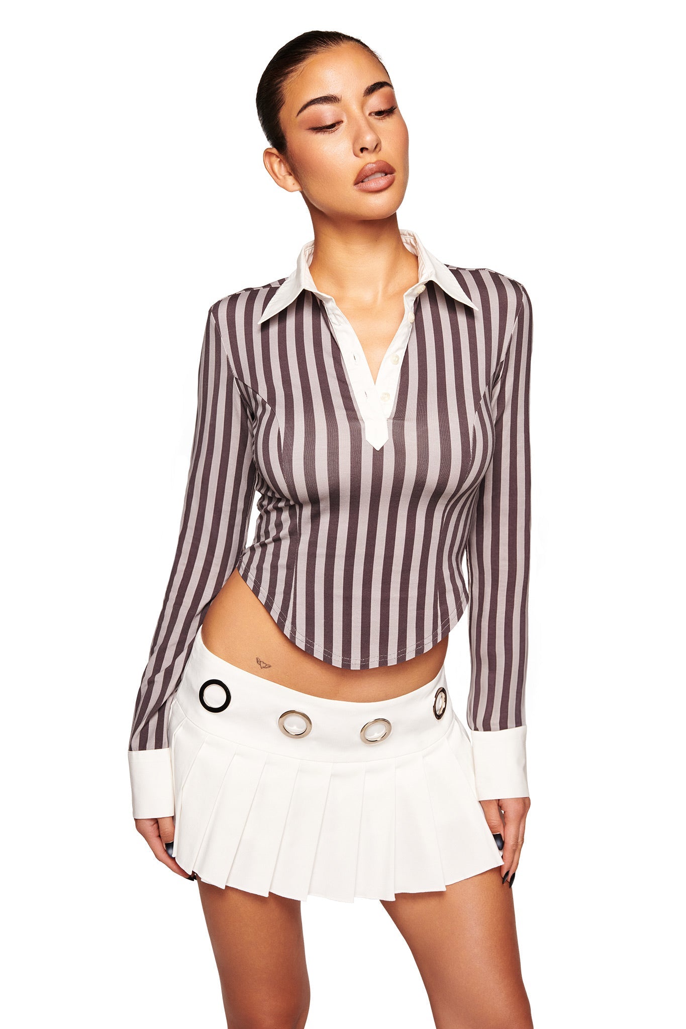 A woman poses against a white background in the I.AM.GIA JULIA TOP - GREY STRIPE, a long-sleeve, bodycon shirt with grey and white vertical stripes and a white collar, paired with a short white pleated skirt with metal grommet waist details.
