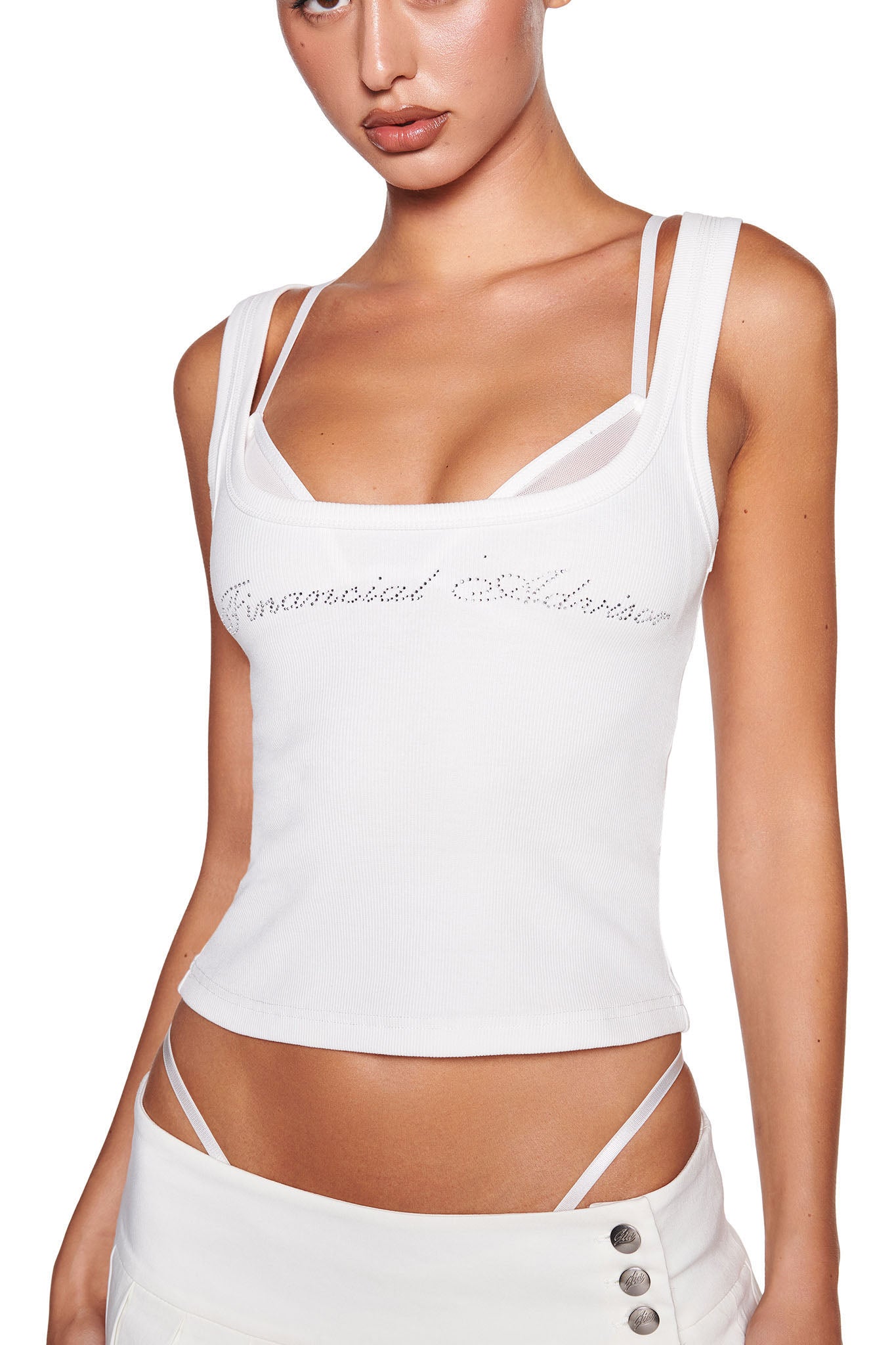 A person is wearing the I.AM.GIA COLE TANK 2.0 - WHITE:DIAMONTE, a white rib knit tank top with thin straps and "Emotional Baggage" spelled out in rhinestones on the front. They are also sporting white low-rise pants featuring visible straps peeking above the waistband and decorative buttons on the side.