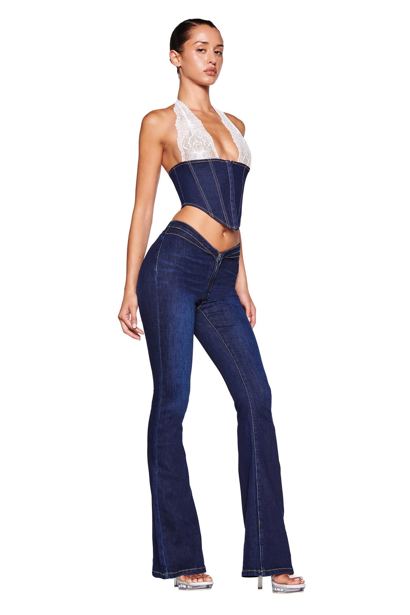 A woman stands modeling a trendy outfit from I.AM.GIA, wearing the ELLENA CORSET - BLUE : DARK WASH. The halter top features a white lace bust and a dark blue corset design, paired with matching high-waisted stretch denim jeans that flare at the bottom. She completes the look with transparent high-heeled sandals.