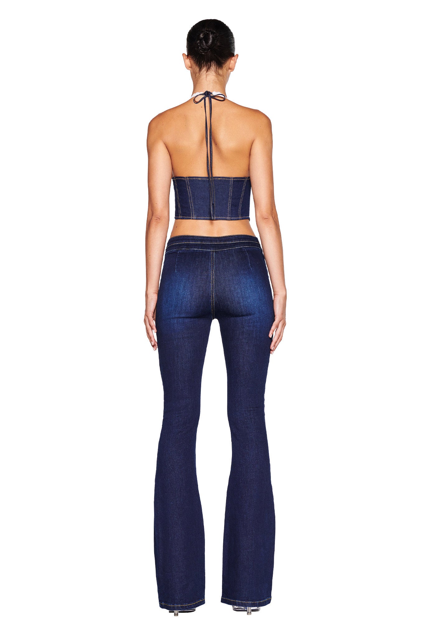 A woman is standing with her back to the camera, showcasing a dark wash denim two-piece outfit from I.AM.GIA. She is wearing the ELLENA CORSET - BLUE, which includes a halter-style crop top and high-waisted, flared pants. Her hair is tied in a neat bun as the image focuses on her back, emphasizing the intricate details of the garment.
