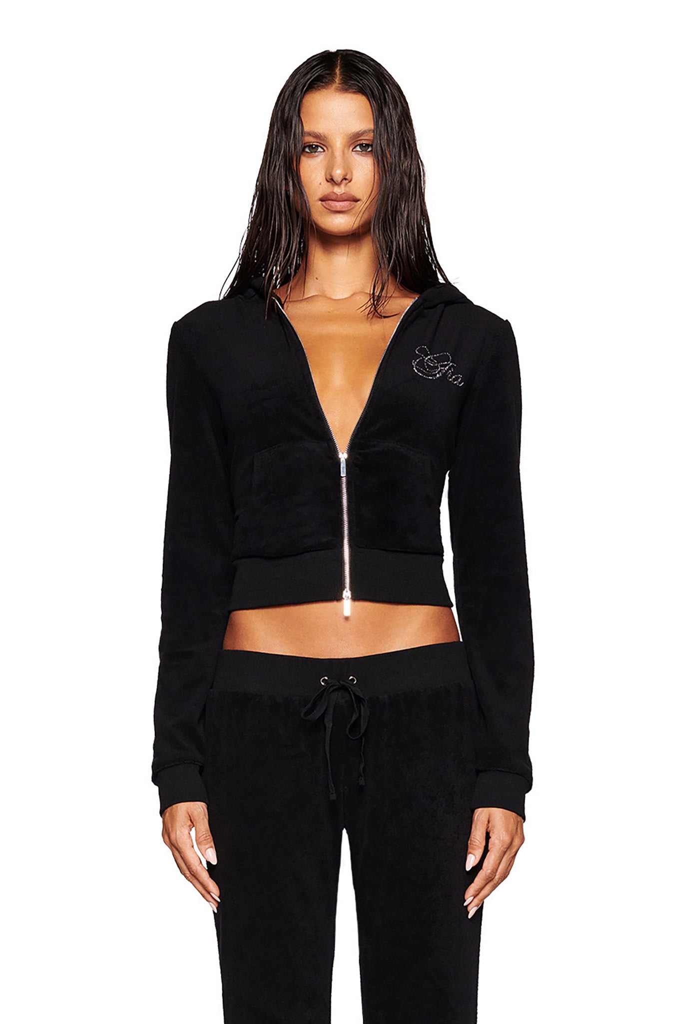 A woman with long wet hair stands facing forward. She is wearing a black KAMERON TERRY JACKET by I.AM.GIA, featuring a small embroidered design on the upper left side, paired with matching black drawstring pants. The jacket is partially unzipped, revealing a bit of her midriff.