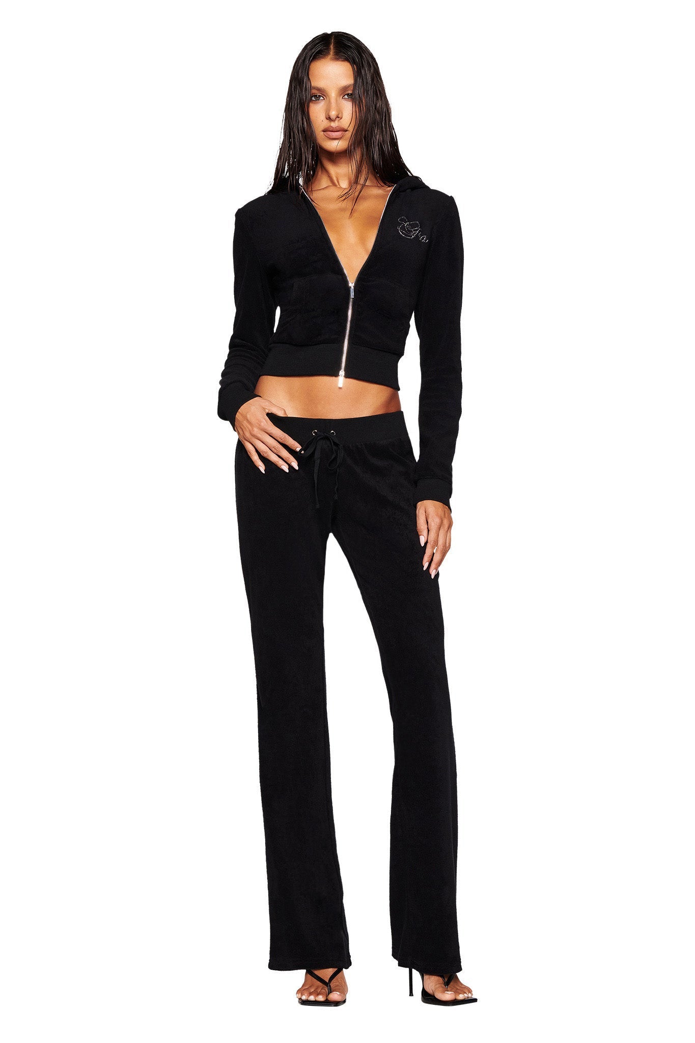 A woman stands against a white background wearing an I.AM.GIA KAMERON TERRY JACKET - BLACK, partially unzipped to reveal a part of her midriff, along with matching black pants. She has long, wet hair and is wearing high-heeled sandals.