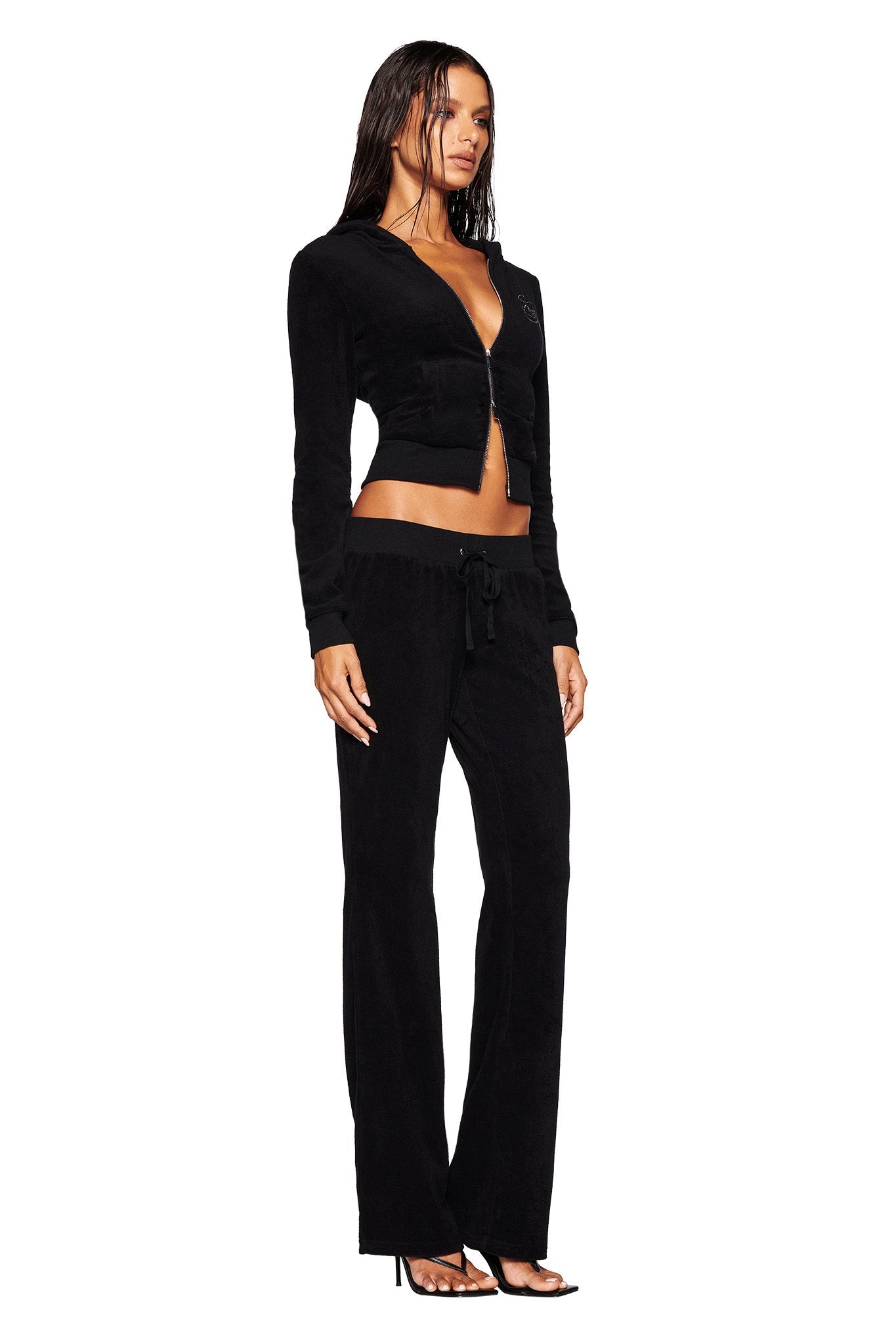 A woman stands wearing a black KAMERON TERRY JACKET - BLACK from I.AM.GIA tracksuit and black heeled sandals. The outfit includes a zip-up hoodie jacket and drawstring pants. She has long, straight hair and a neutral expression, posing against a white background.