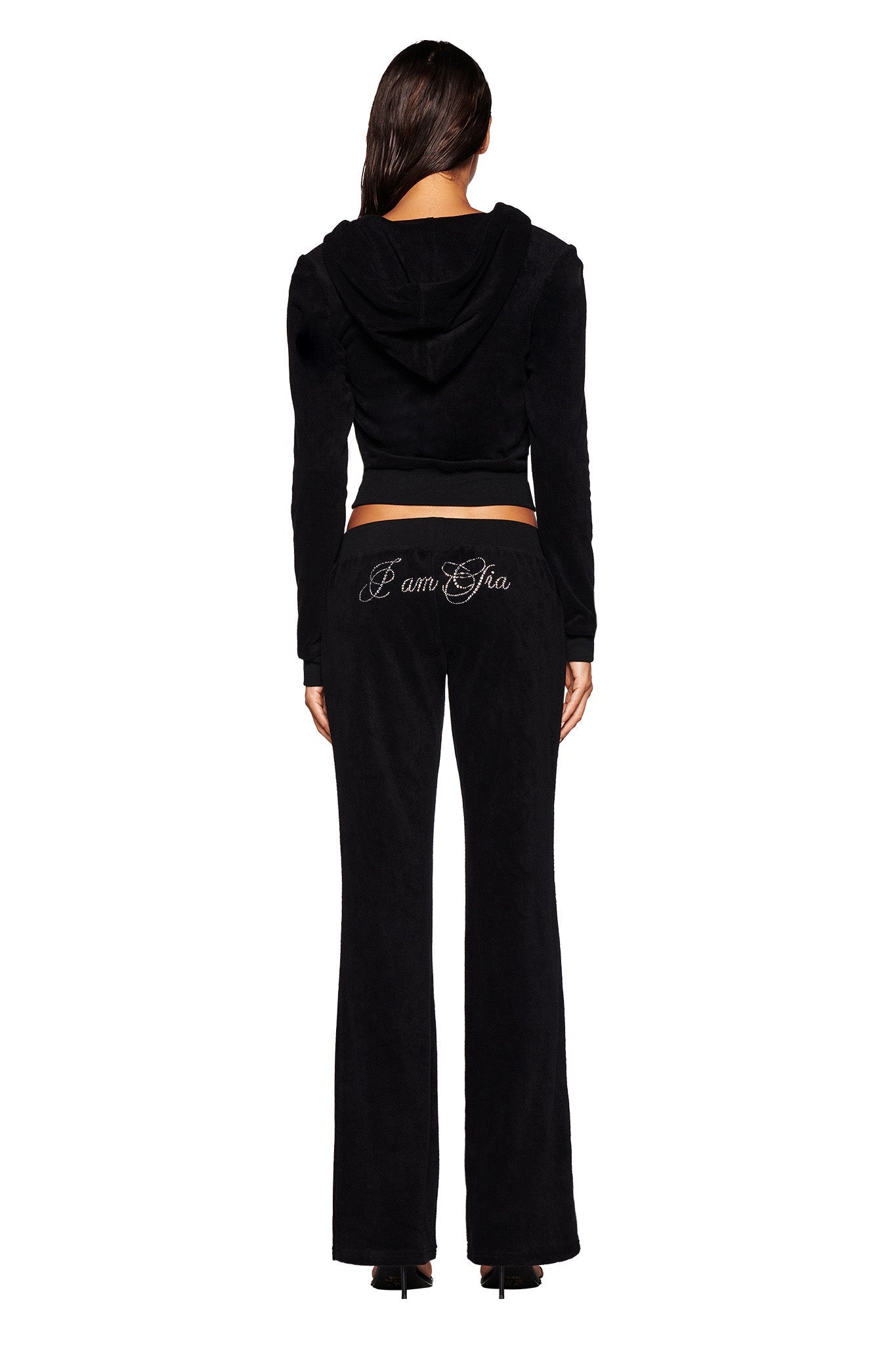 A woman is standing with her back to the camera, wearing a stylish black KAMERON TERRY JACKET from I.AM.GIA. The hooded jacket and matching pants feature a custom print, with "I Am Gia" written in cursive across the back of the pants, making the outfit both chic and comfortable.