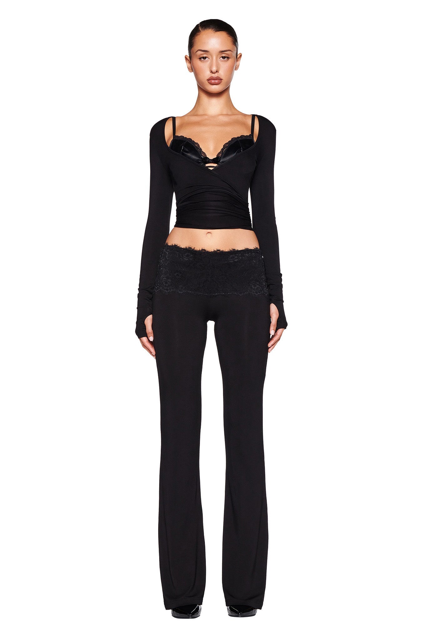 A person with long hair tied back is standing against a white background. They are wearing the MANDY TOP - BLACK from I.AM.GIA, which is a black wrap top made from super soft stretch bamboo with lace details around the neckline, paired with black pants featuring lace trim at the waist. Their hands are relaxed at their sides.