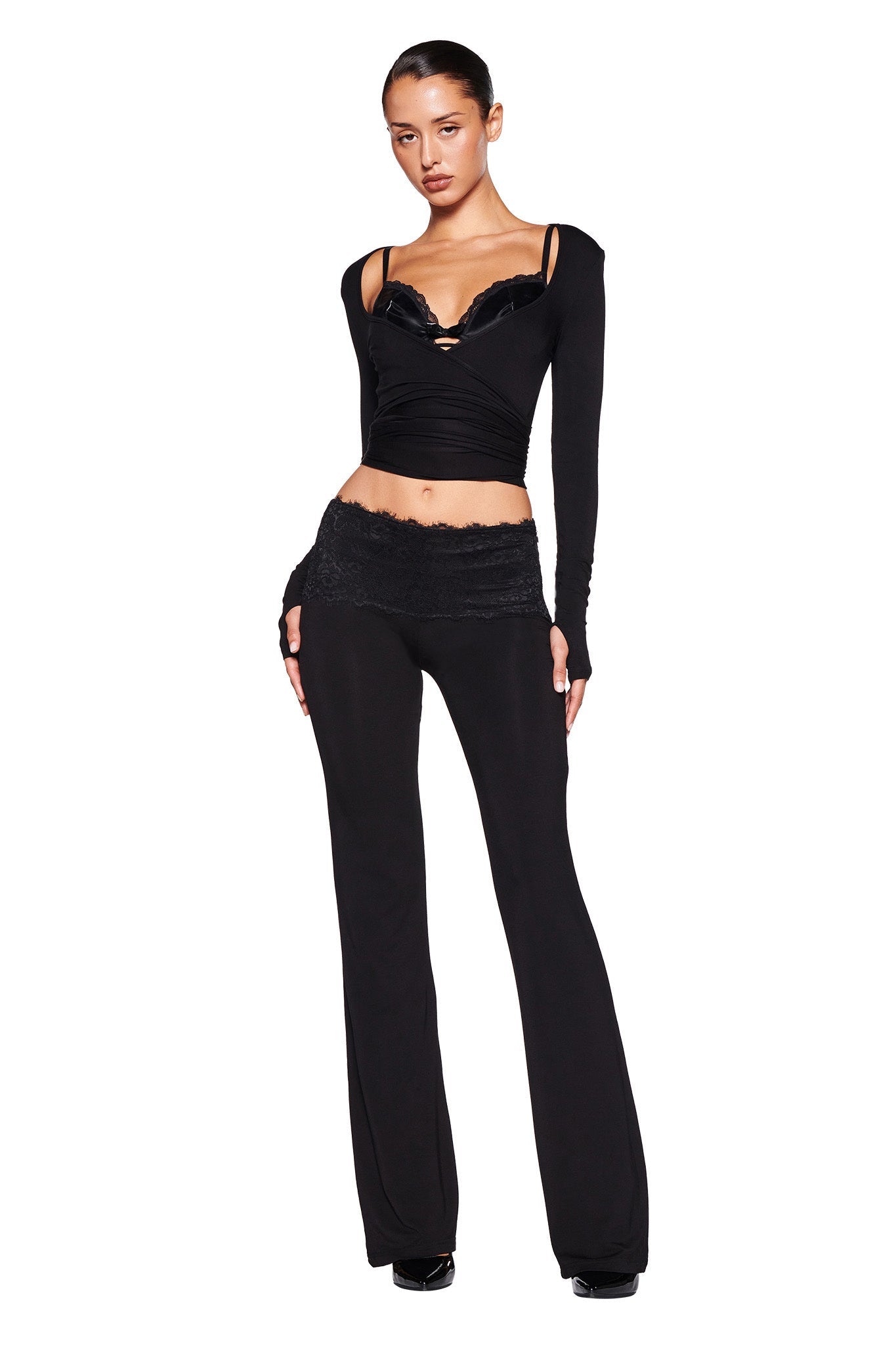 A person stands in a confident pose wearing a stylish all-black outfit from I.AM.GIA. The bodycon silhouette includes the MANDY TOP - BLACK, featuring long sleeves and a fitted design with lace trim on the neckline, paired with flared pants that have lace detailing at the waist. The background is plain white.