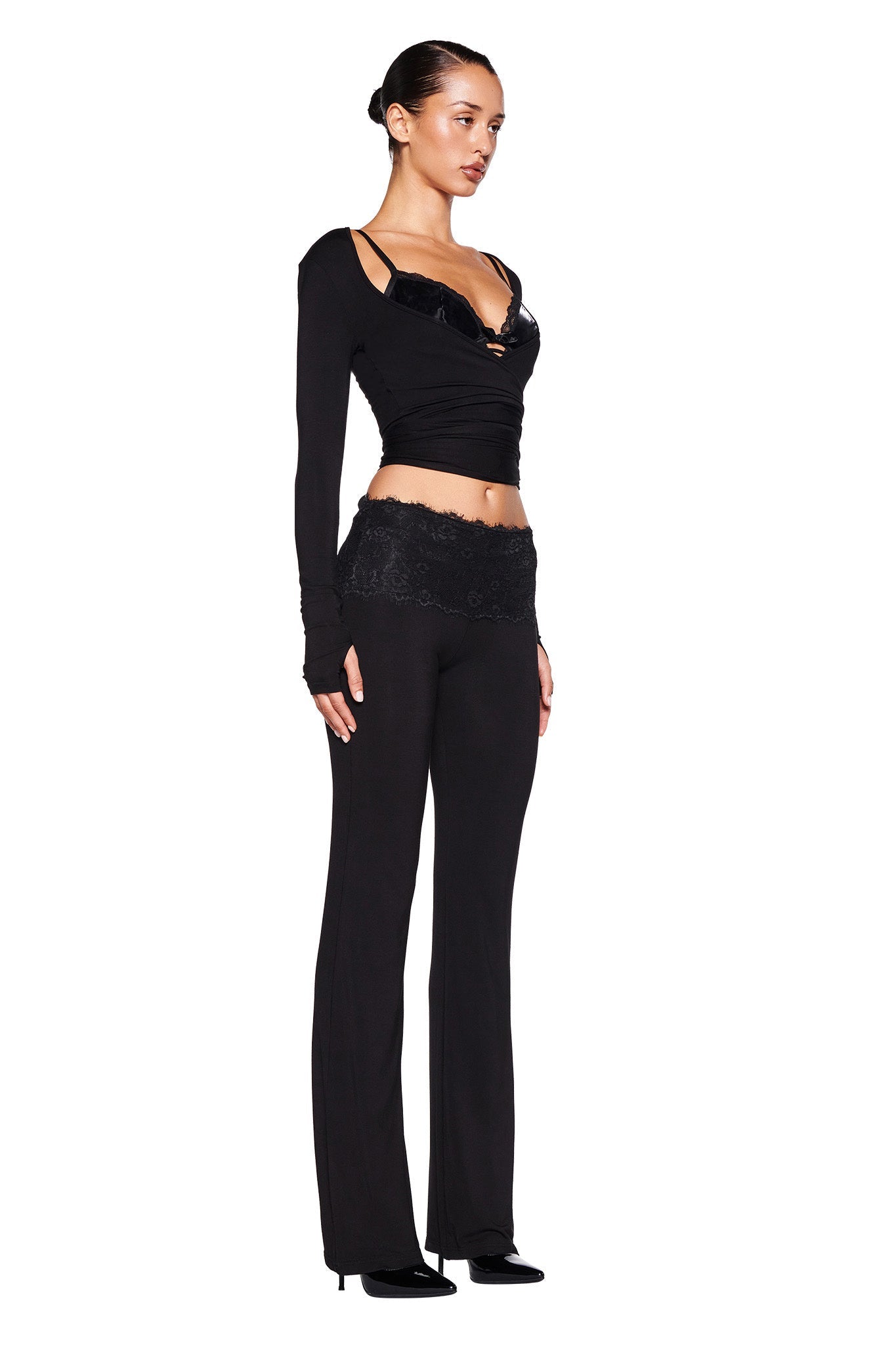 A woman stands against a plain white background, facing slightly to the left. She is wearing the MANDY TOP - BLACK by I.AM.GIA, featuring a bodycon silhouette and black high-waisted pants with a lace waistband. Her hair is slicked back into a bun, and she is wearing black high-heeled shoes.