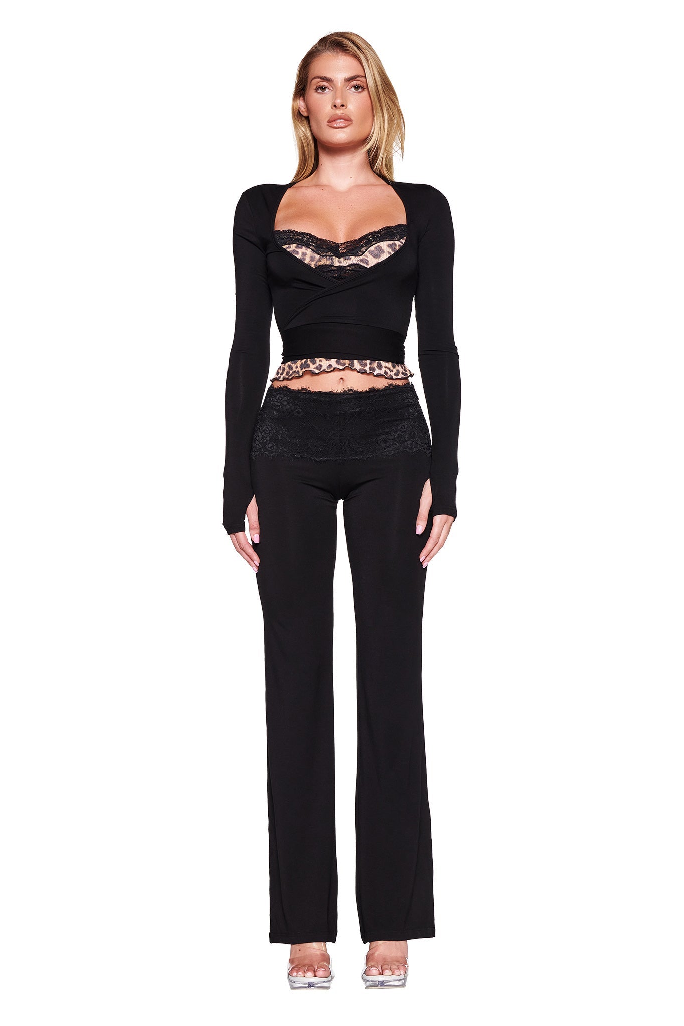 A woman stands against a white background, modeling the MANDY TOP - BLACK by I.AM.GIA. This fitted black wrap top features a revealing neckline and intricate lace detailing, crafted from super soft stretch bamboo fabric that perfectly accentuates her bodycon silhouette. She complements the top with matching black pants and clear high heels. Her hair is styled straight, and she gazes forward with a neutral expression.