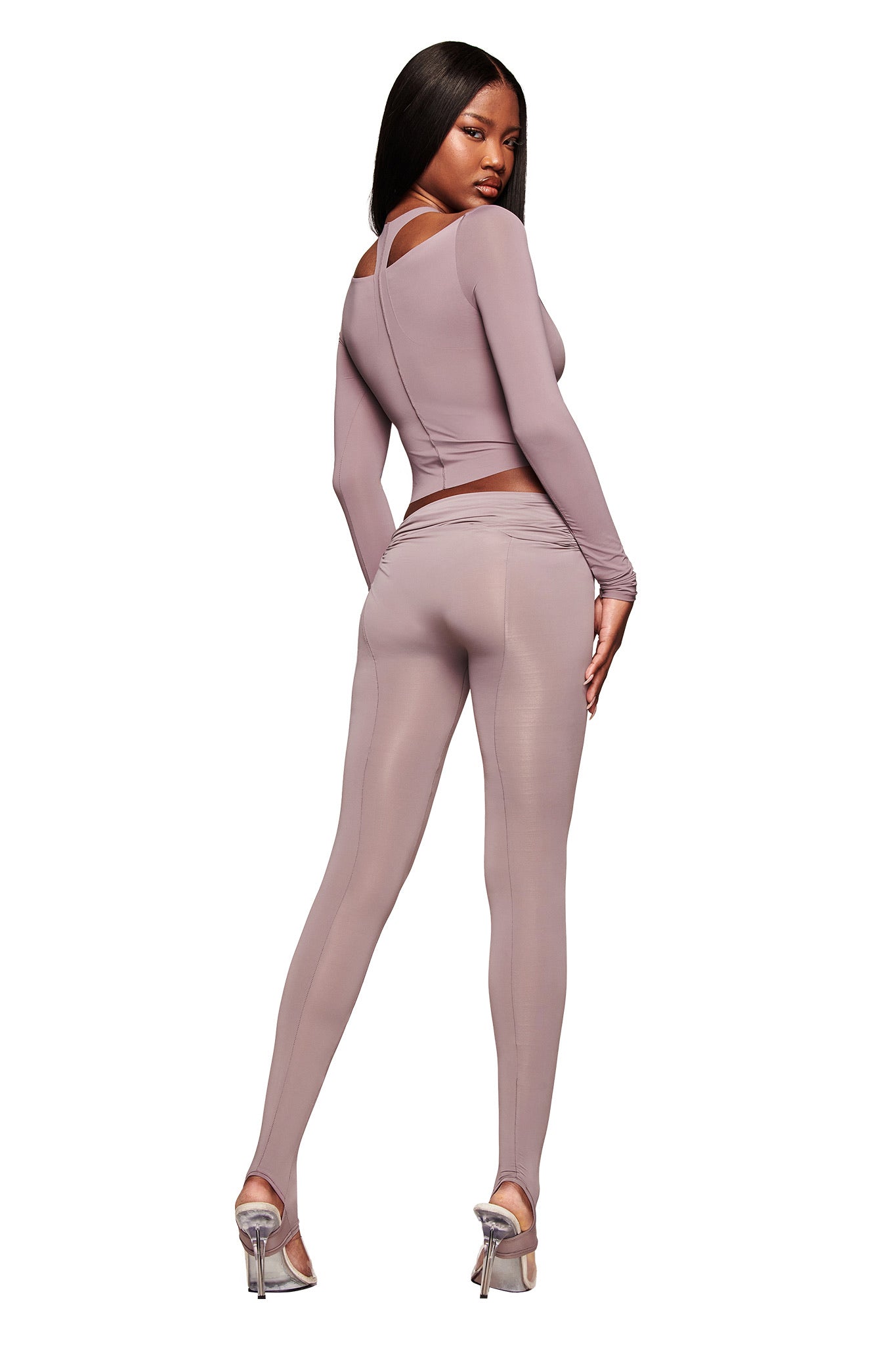 A woman with long dark hair poses with her back to the camera, wearing the I.AM.GIA NERINA LONG SLEEVE TOP in dusty lilac and matching leggings, paired with silver high heels against a plain white background.