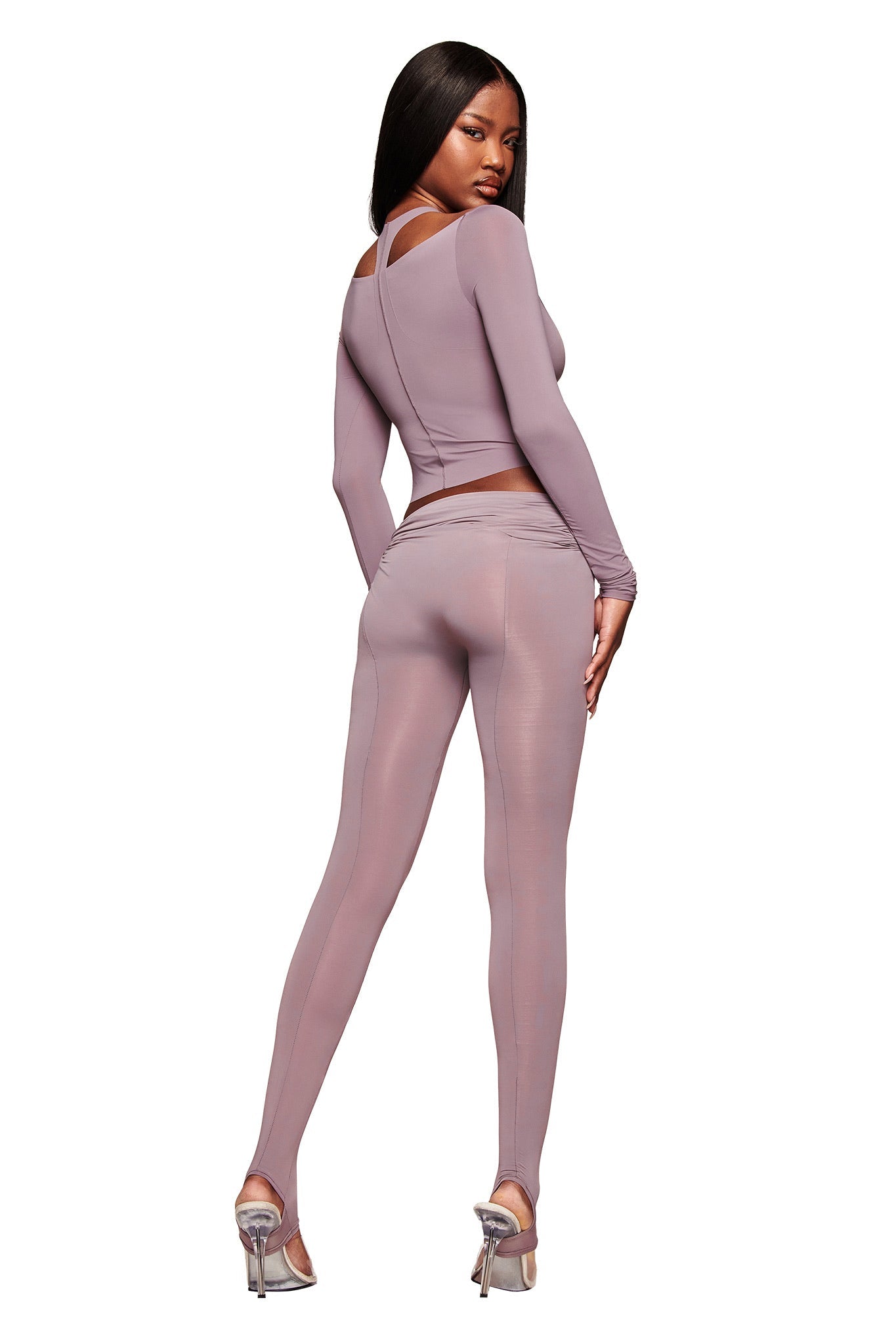A woman with long, straight hair poses against a white background in the I.AM.GIA NERINA LEGGING in dusty lilac and a matching fitted long-sleeve top, styled with silver high-heeled shoes.