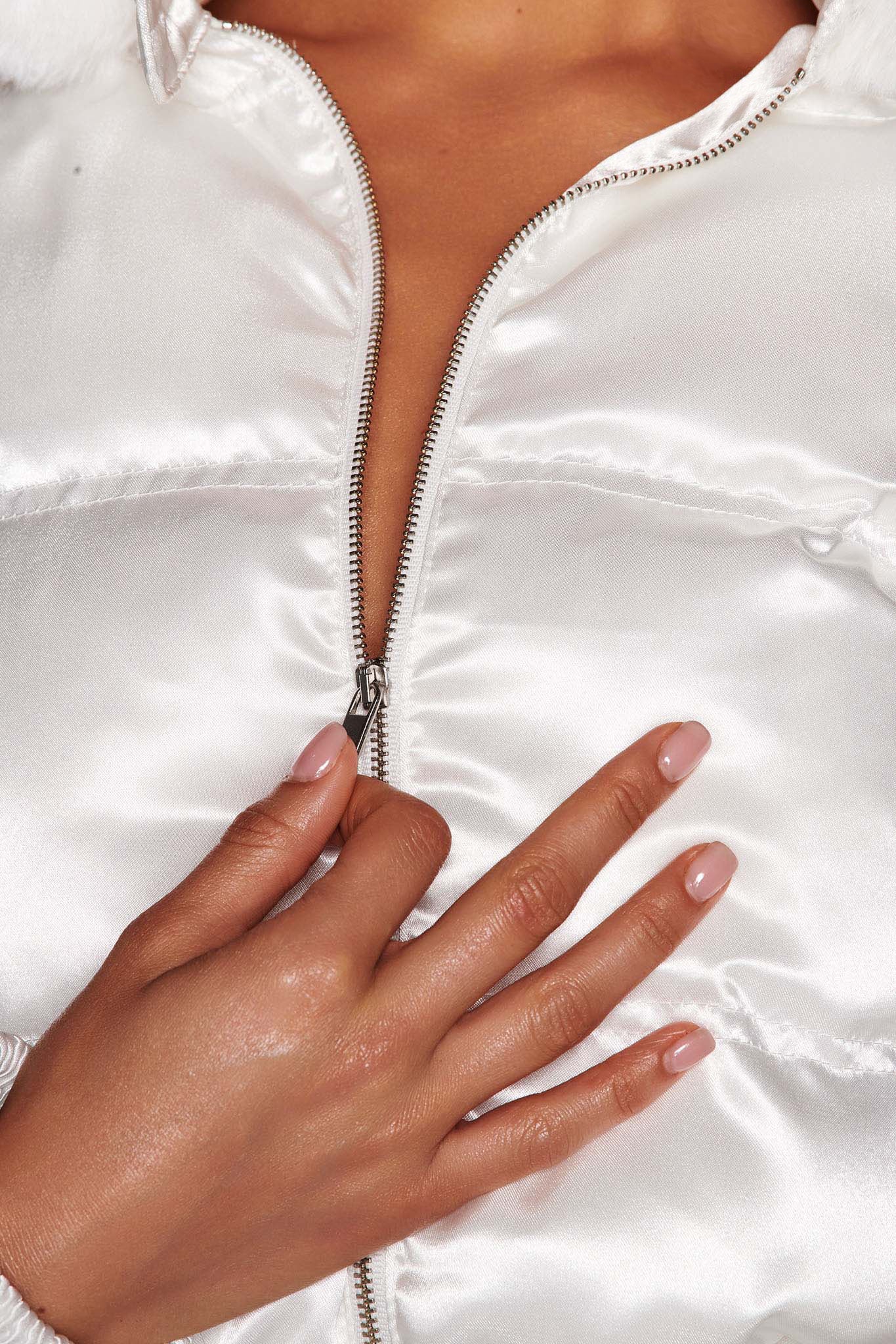 Close-up of a person wearing the shiny white RUBIA JACKET 2.0 from I.AM.GIA, partially zipped. Their hand, adorned with light pink nail polish, rests near the zipper, accentuating the glossy texture of the satin fabrication and their elegant manicure.