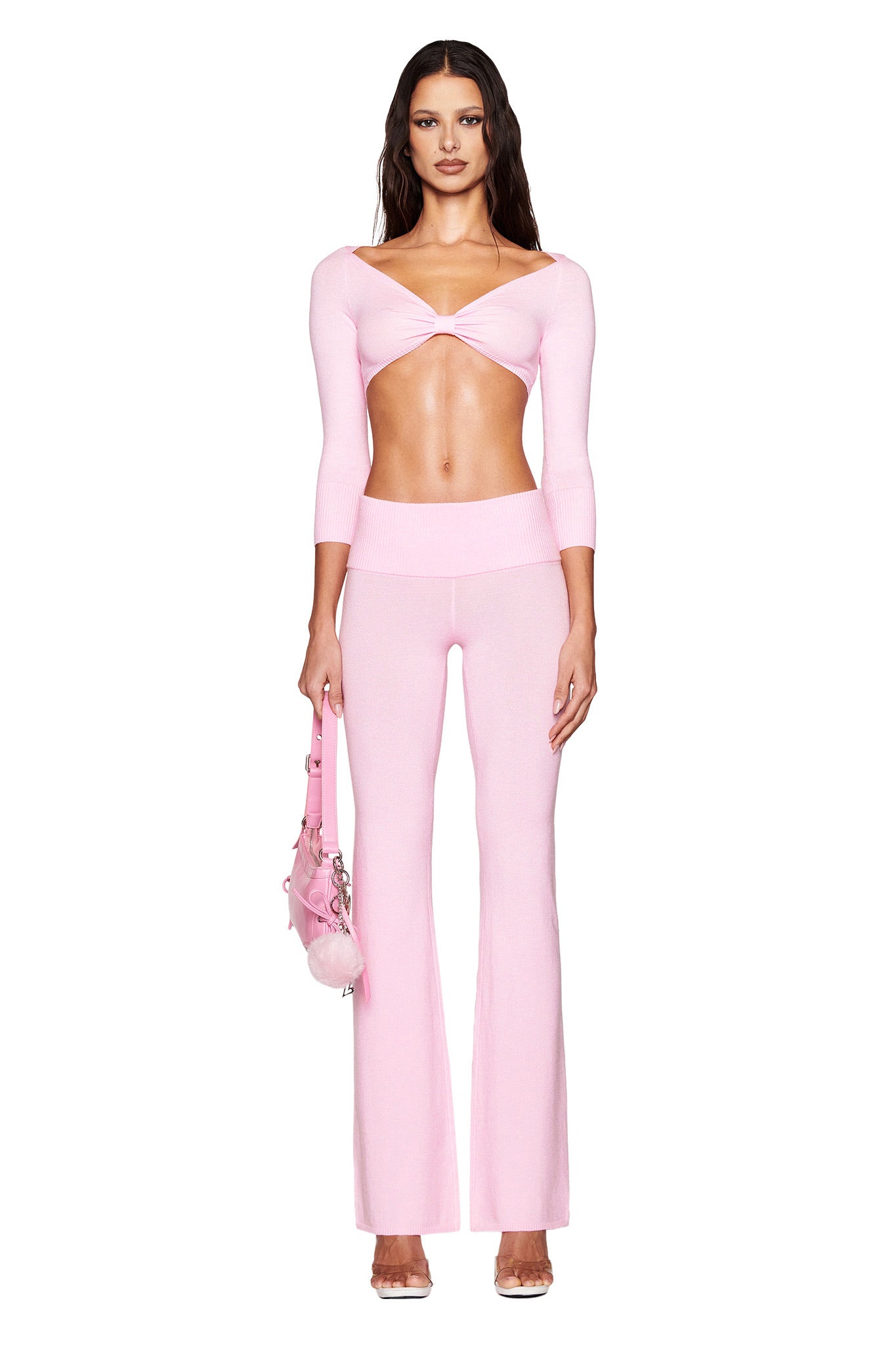 A woman showcases I.AM.GIA's MORGANA PANT in baby pink, featuring a form-fitting ultra-soft knit crop top with a twist front and matching flare pants. She holds an embellished pink bag, her long dark hair draping over her shoulders against a plain white background.