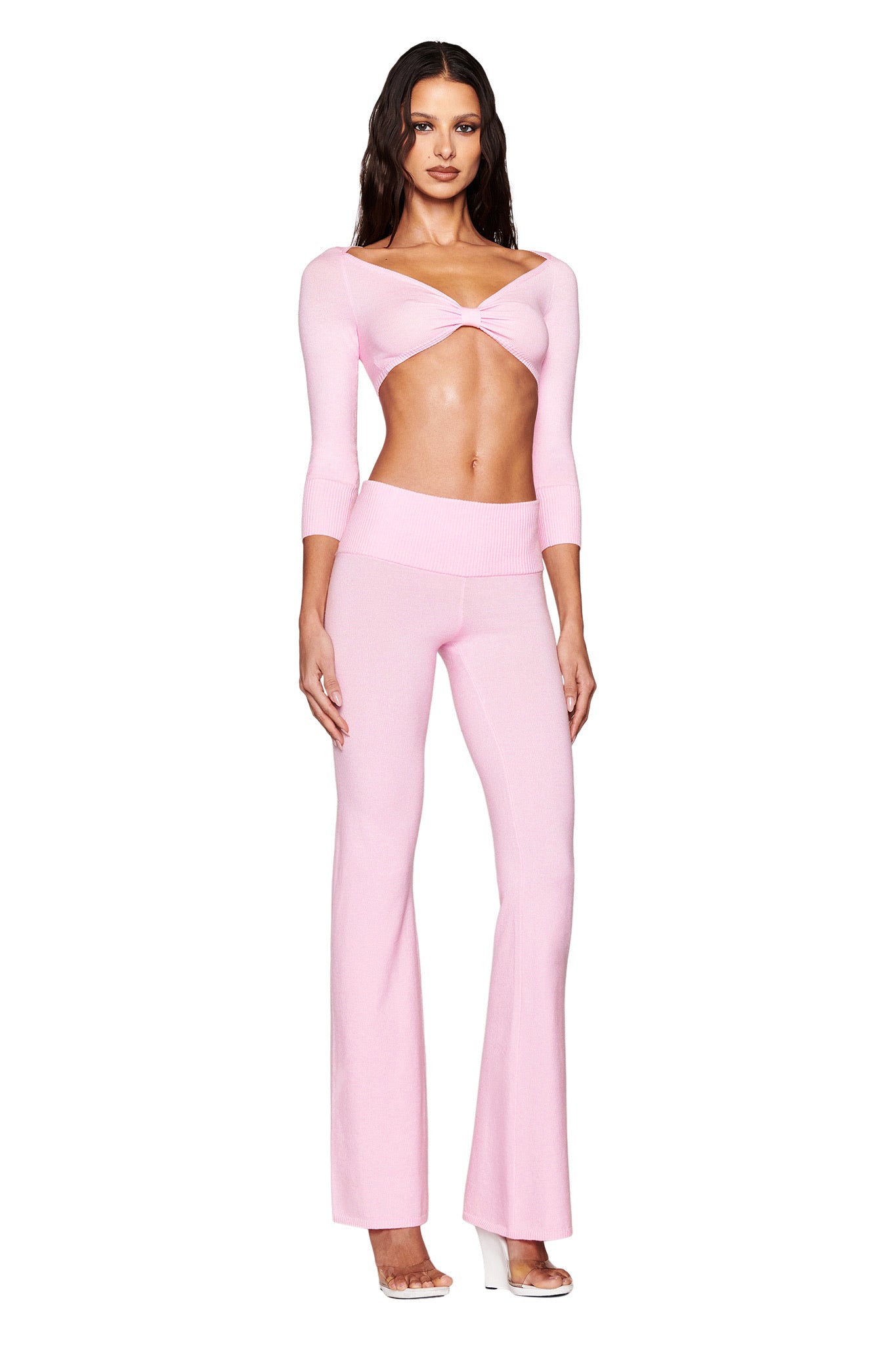 Someone poses confidently in an I.AM.GIA MORGANA CROP TOP - PINK, characterized by its twisted front detail. Paired with matching high-waisted, flared pants and clear heels, their long wavy hair adds to the effortless style against a white backdrop.