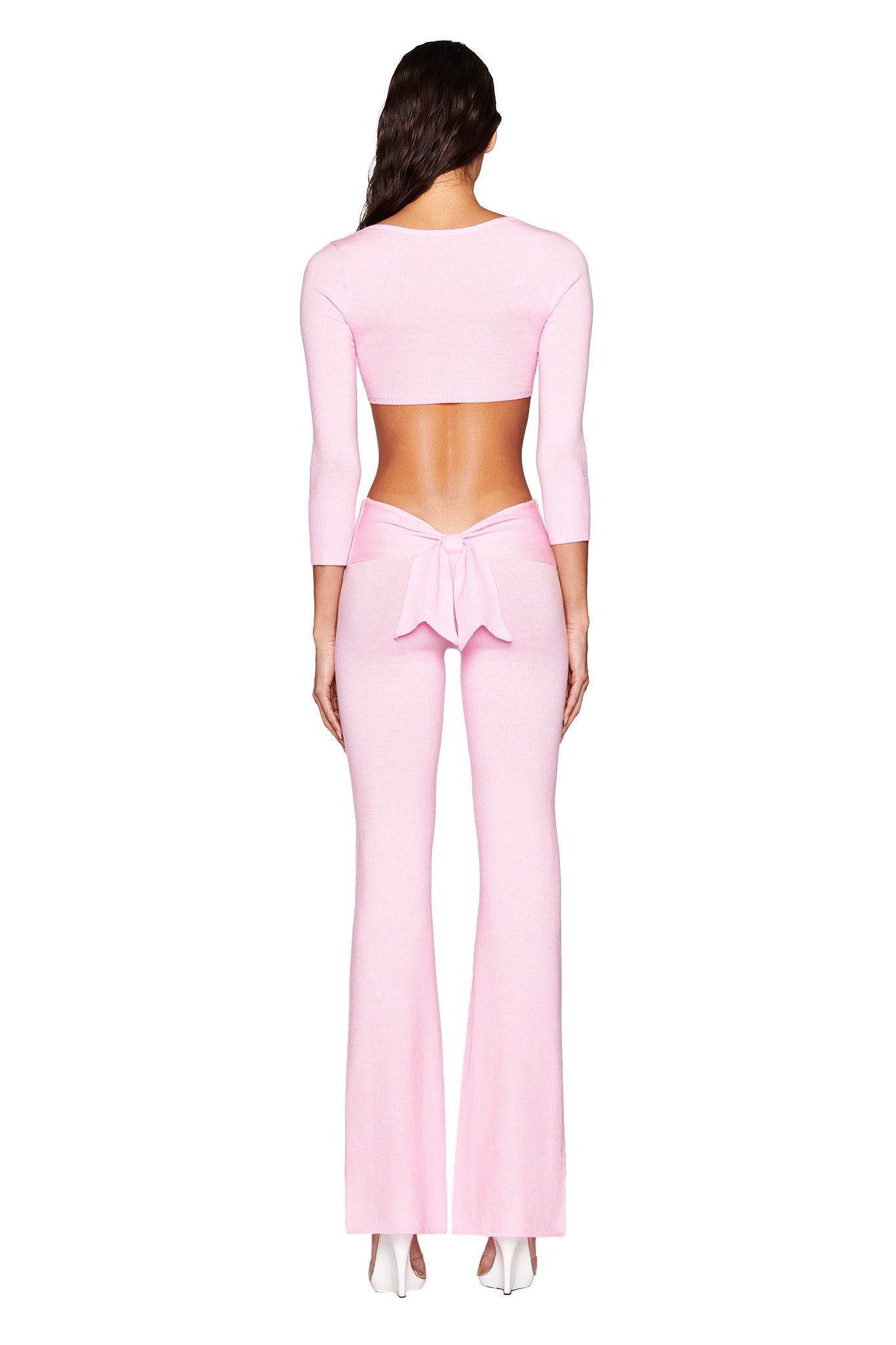 Wearing I.AM.GIA's MORGANA CROP TOP in pink, paired with form-fitting flared pants tied with a bow, this person stands back to the camera in ultra-soft knit fabric and white high-heeled shoes, highlighting the plunging neckline and super-cropped style.