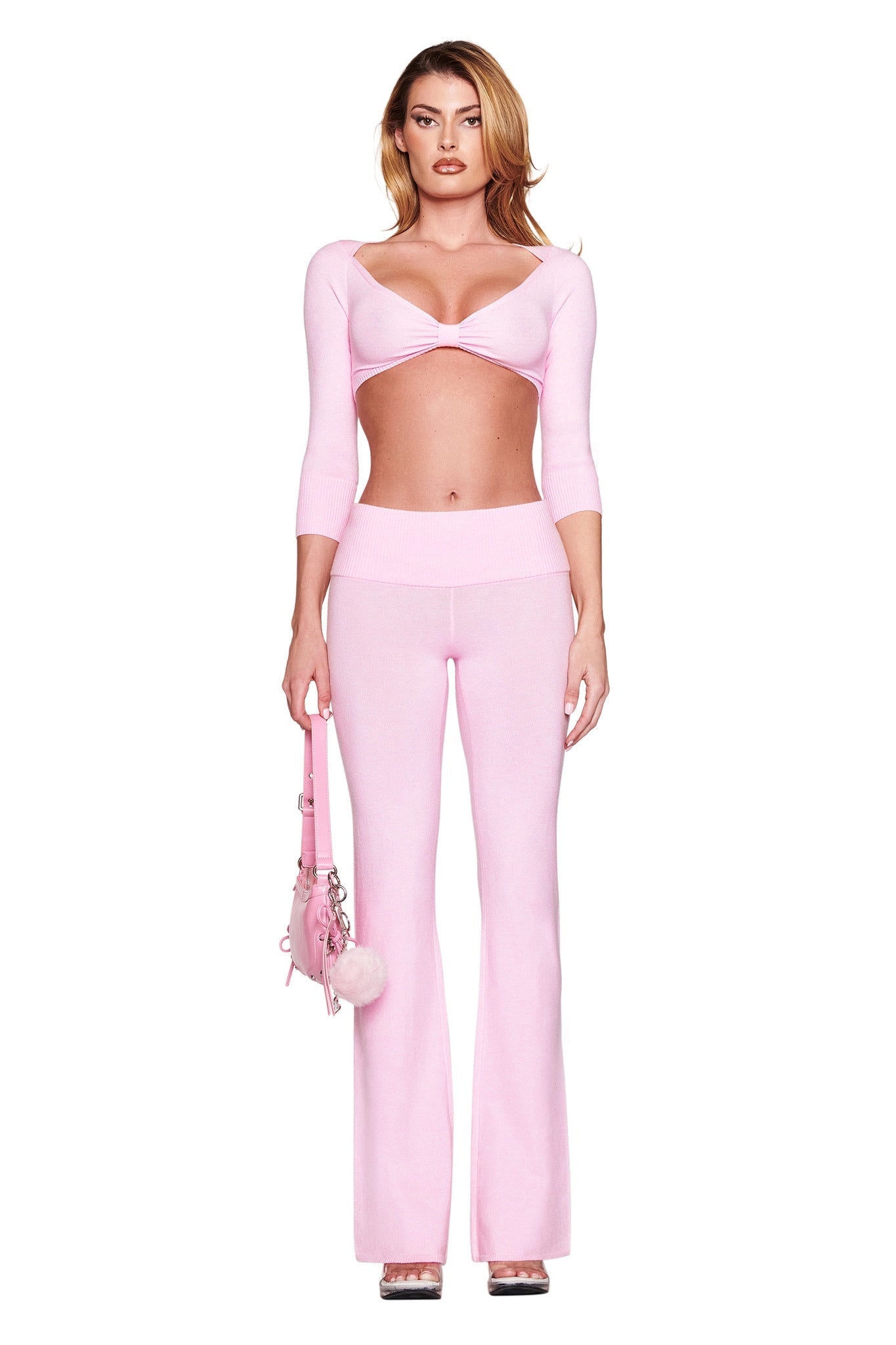A person in the MORGANA CROP TOP - PINK by I.AM.GIA with flared pants holds a pink handbag adorned with fluffy pom poms, standing gracefully against a plain white background and exuding style and charm.