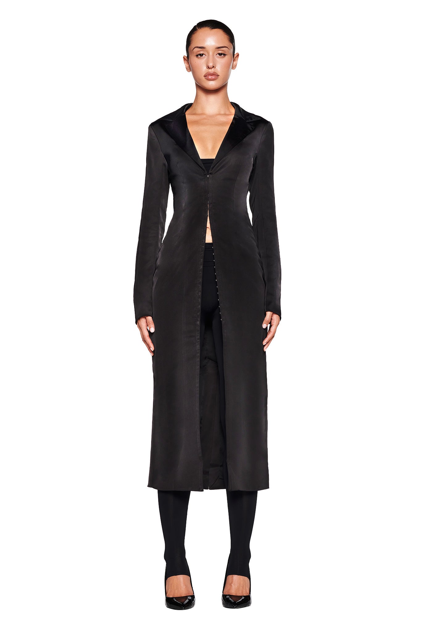A person stands against a white background, wearing the I.AM.GIA RAENA COAT in black with a deep V-neck over black pants and black shoes. The long, fitted coat, made from satin fabrication and featuring a tailored fit, is buttoned at the waist and has long sleeves. Their hair is pulled back.