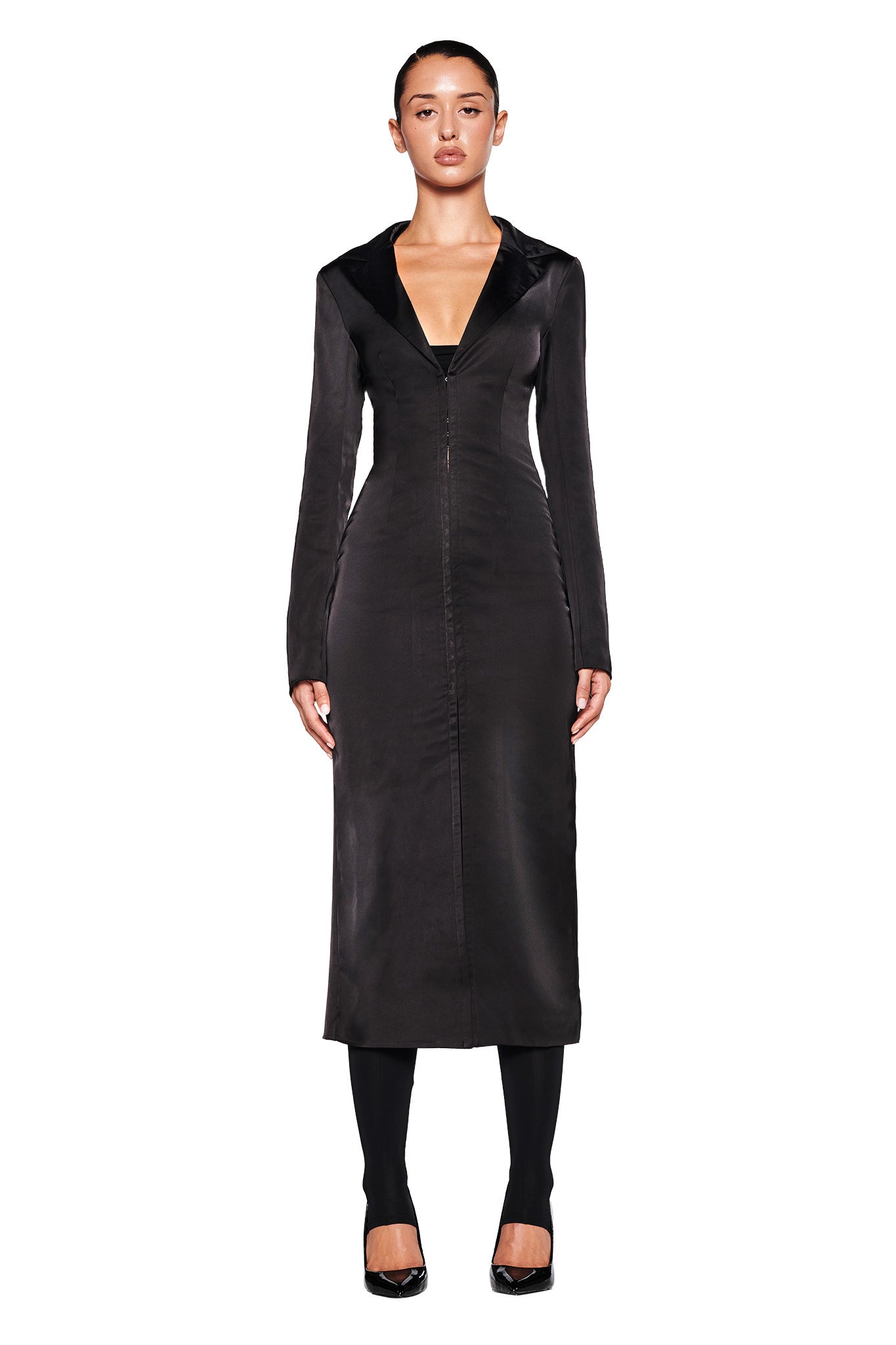 A person stands facing forward wearing the RAENA COAT - BLACK from I.AM.GIA. The satin fabrication of the coat adds a touch of elegance as it falls below the knees. They are also wearing black leggings and shoes, with their hair pulled back neatly. The background is plain white.