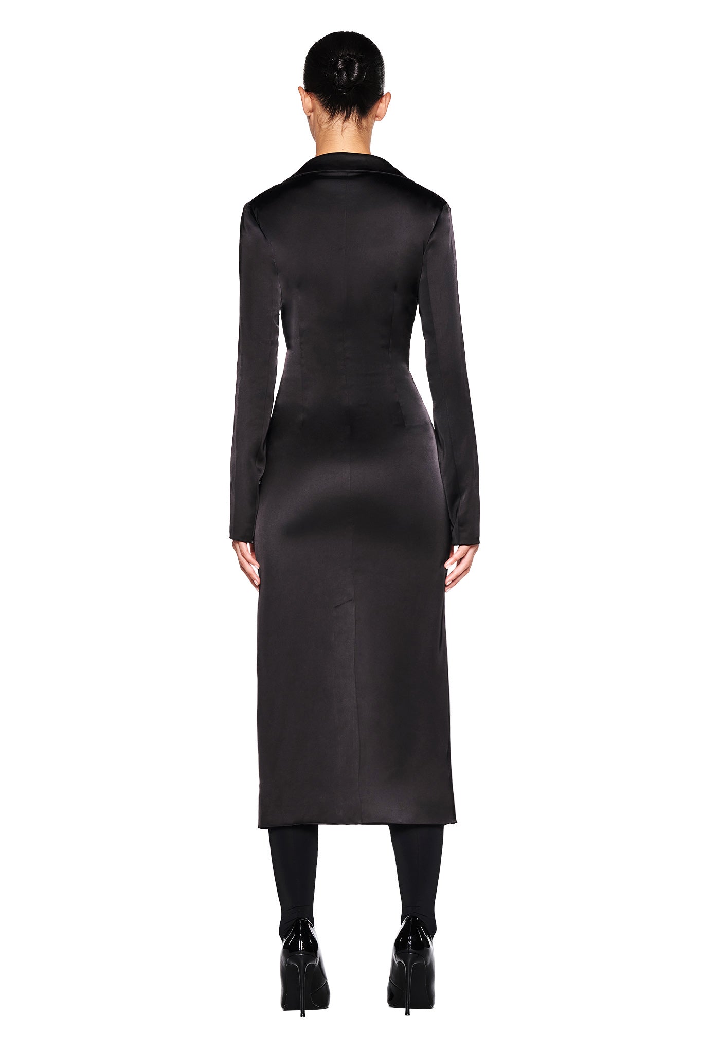 A person with their hair in a bun is seen from behind, wearing the "RAENA COAT - BLACK" by I.AM.GIA. The long, satin fabrication coat has a tailored fit that extends to mid-calf. They also wear black stockings and black high-heeled shoes against a white background.