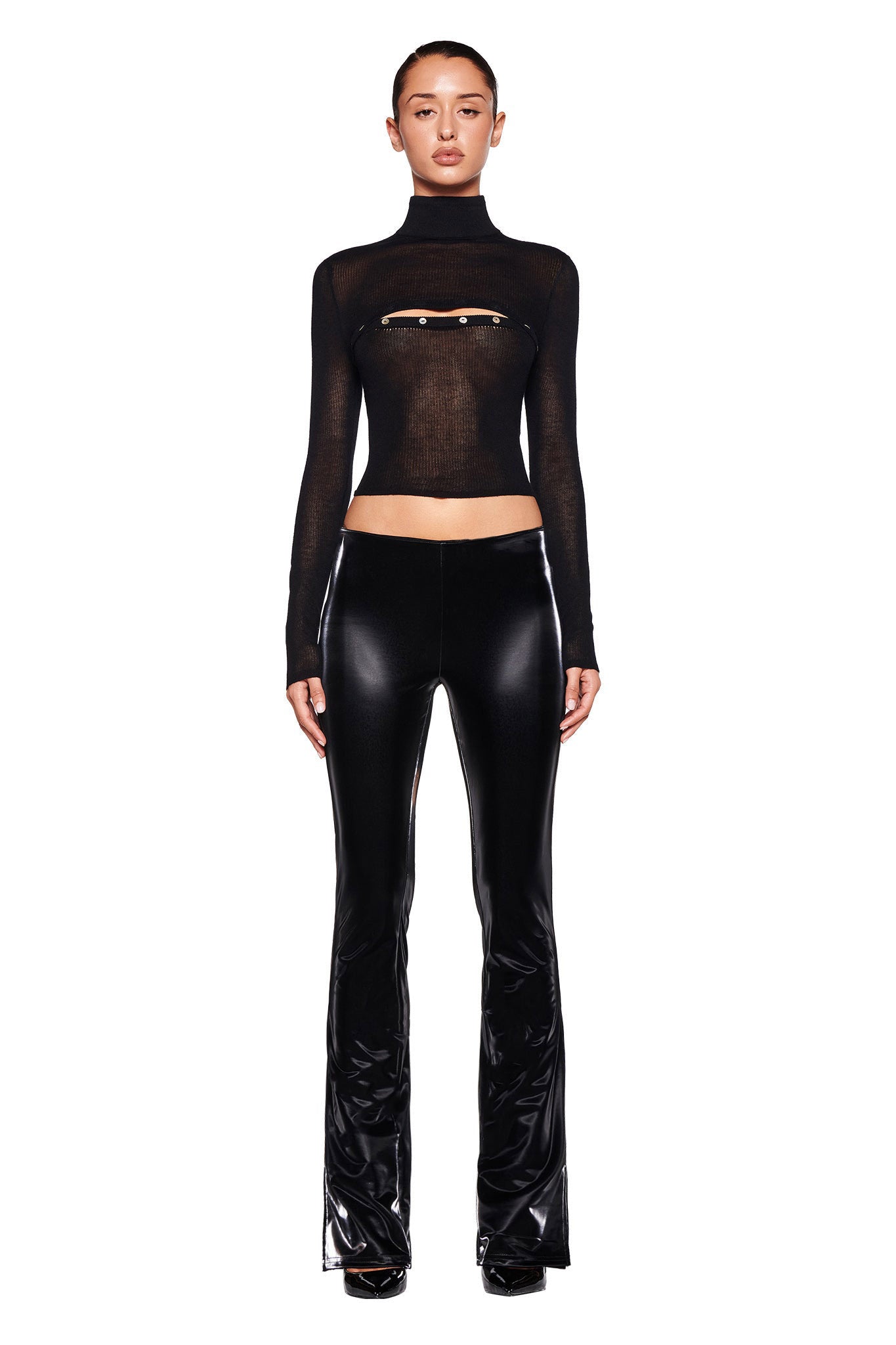 A person stands against a white background wearing the STORM TOP in black by I.AM.GIA, featuring long sleeves and a semi-sheer design with a small horizontal zipper opening at the chest. They pair it with bodycon fit shiny black flare pants, have sleek hair pulled back, and wear black high-heeled shoes.
