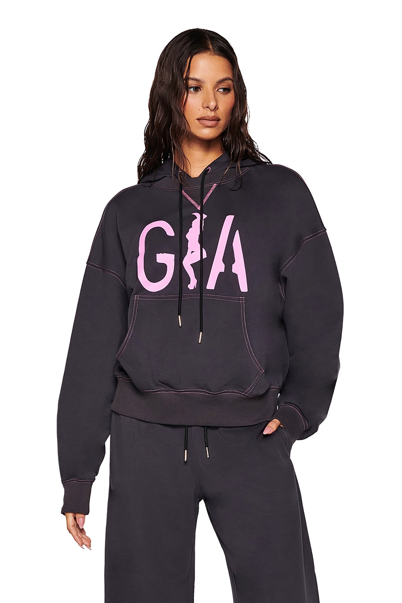 A person in an oversized fit SPENCER HOODIE - GREY from I.AM.GIA, featuring 'GIA' artwork in pink, stands with hands by their sides. The charcoal grey hoodie includes a large pocket and drawstring. They are wearing matching pants and have long dark hair against a plain white background.