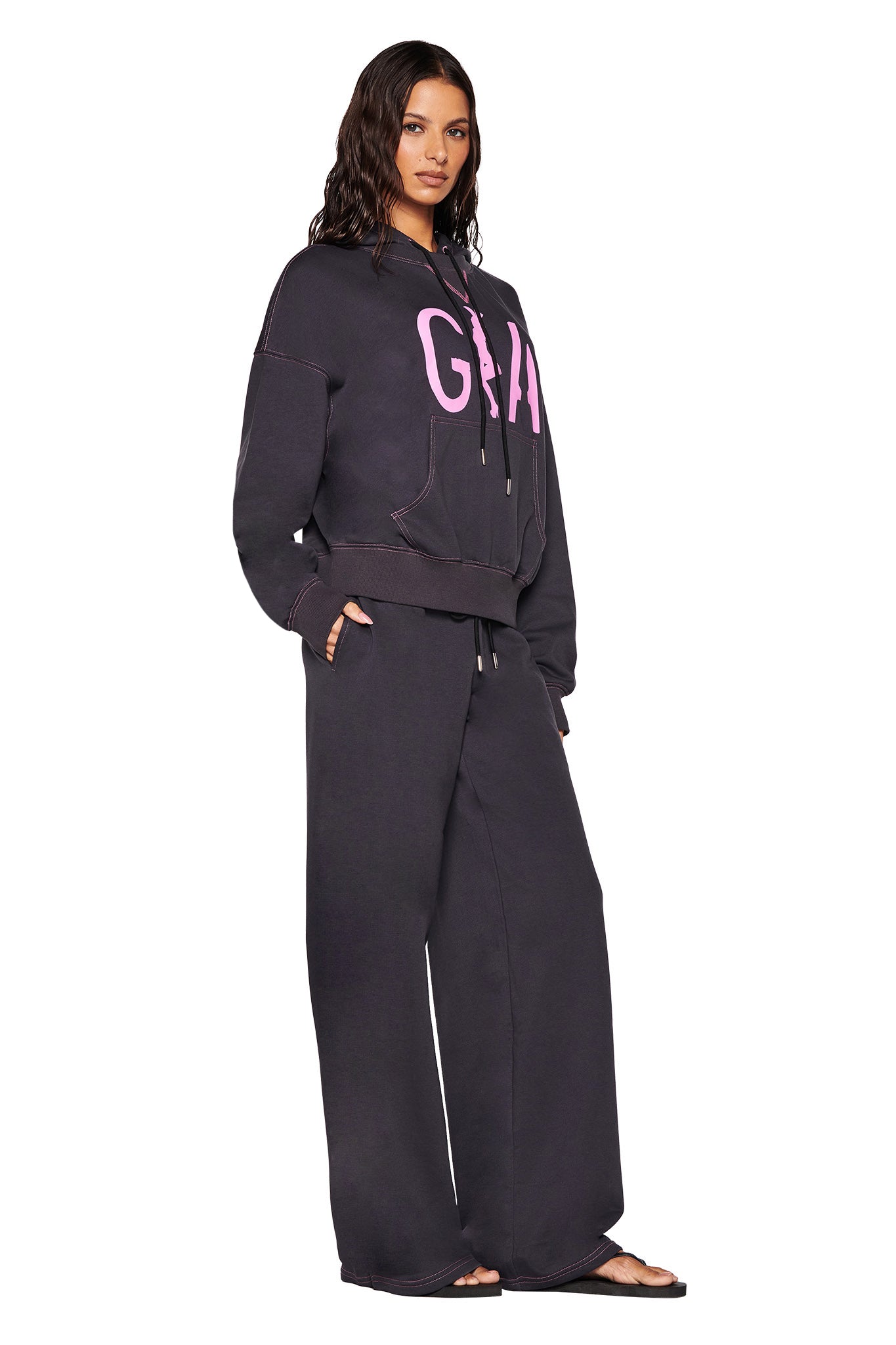 A person is wearing an oversized fit SPENCER HOODIE in charcoal grey by I.AM.GIA, featuring pink 'GIA' artwork. They pair it with matching wide-leg pants and complete the look with black flip-flops while standing against a white background.