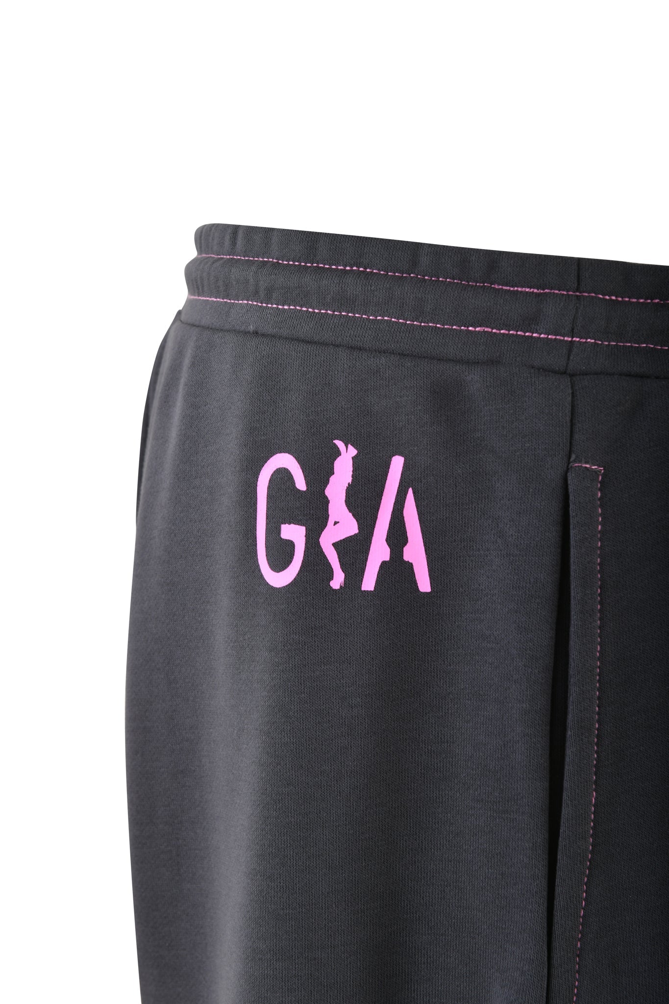 A close-up of the SPENCER PANT in charcoal grey from I.AM.GIA showcases a distinct GIA print along the side. The pink logo displays the letters "GA" with a stylized silhouette of a person integrated between them, while hot pink contrast stitching highlights the garment's edges.