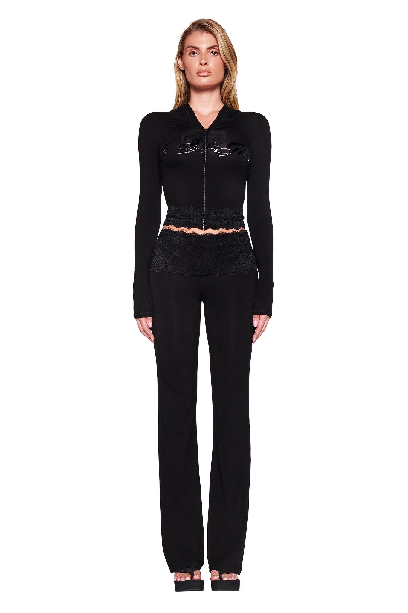 A woman stands facing forward against a white background. She is wearing the TARSHA JACKET - BLACK : TRUE BLACK by I.AM.GIA, which features lace and shiny material elements and showcases a bodycon silhouette. She pairs it with black pants. Her long, wavy blonde hair cascades over her shoulders, and she completes the look with black heeled shoes.
