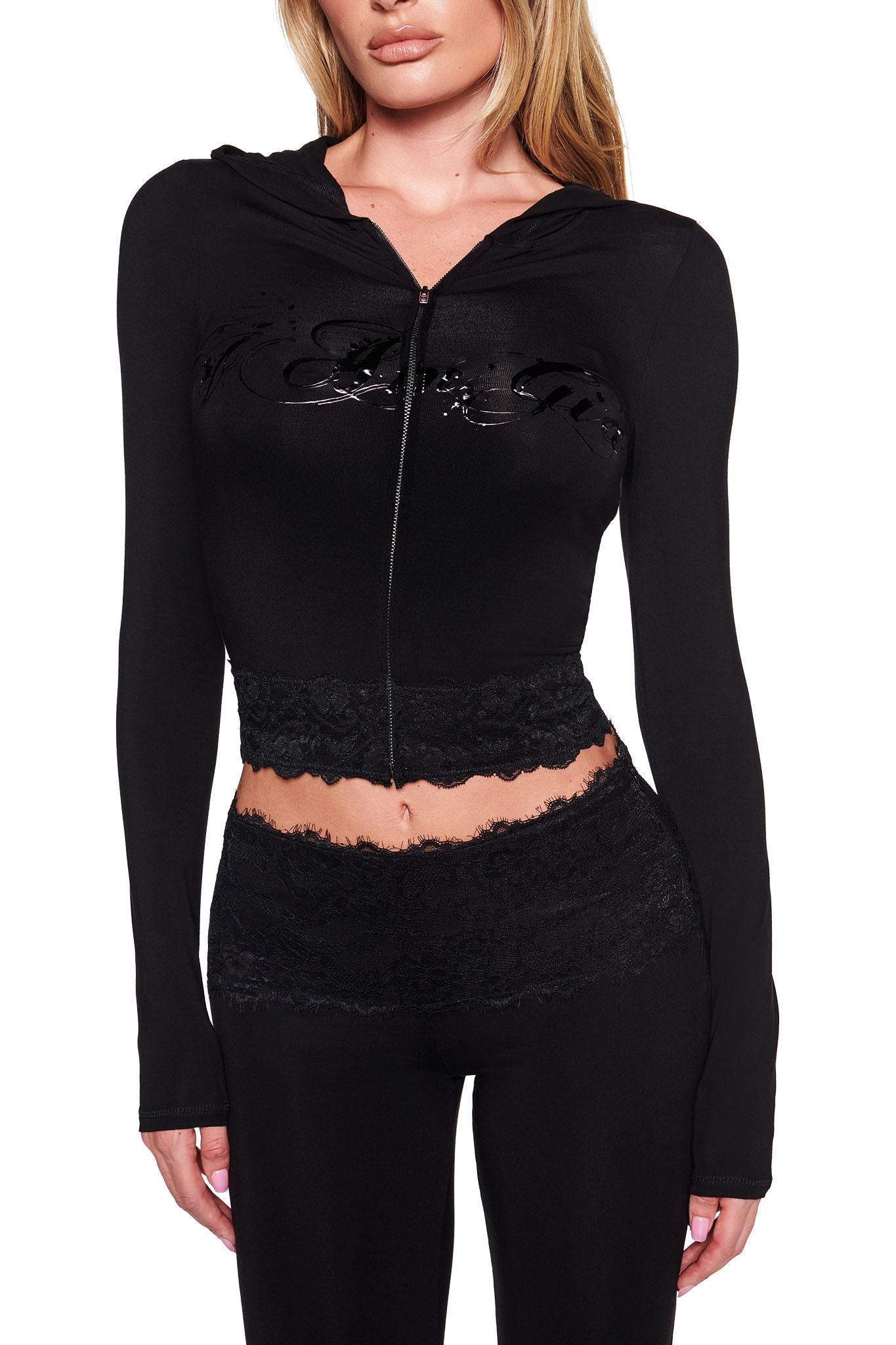 A person is wearing a form-fitting black outfit consisting of the TARSHA JACKET in True Black by I.AM.GIA paired with matching black pants. The jacket, which has a bodycon silhouette, features visible lace trims at the bottom and waistband, and showcases the I.AM.GIA logo on its chest.