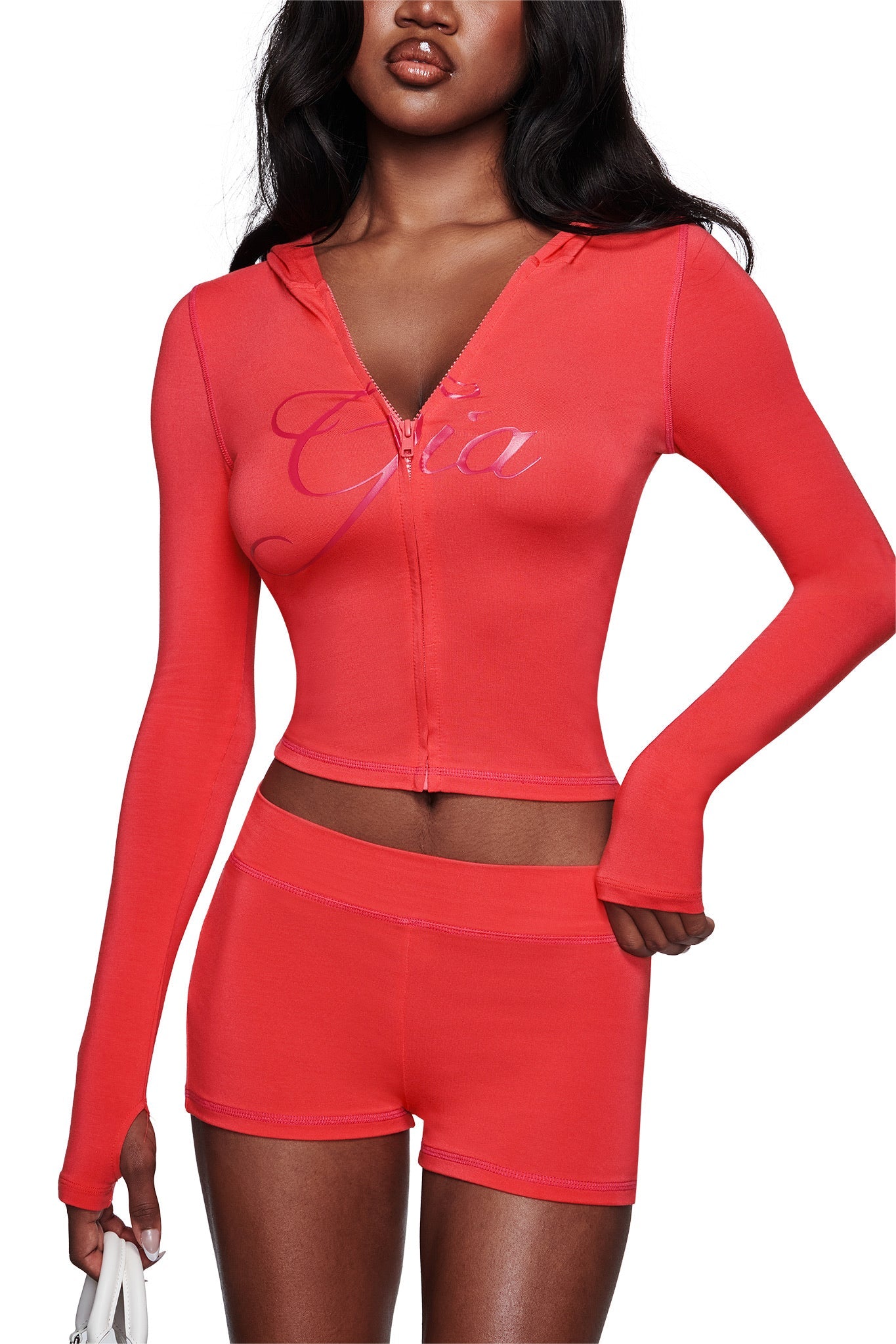 A woman wears the I.AM.GIA BLARE SHORT - PAPAYA and a matching pink zip-up cropped hoodie, holding a small white handbag against a white background.
