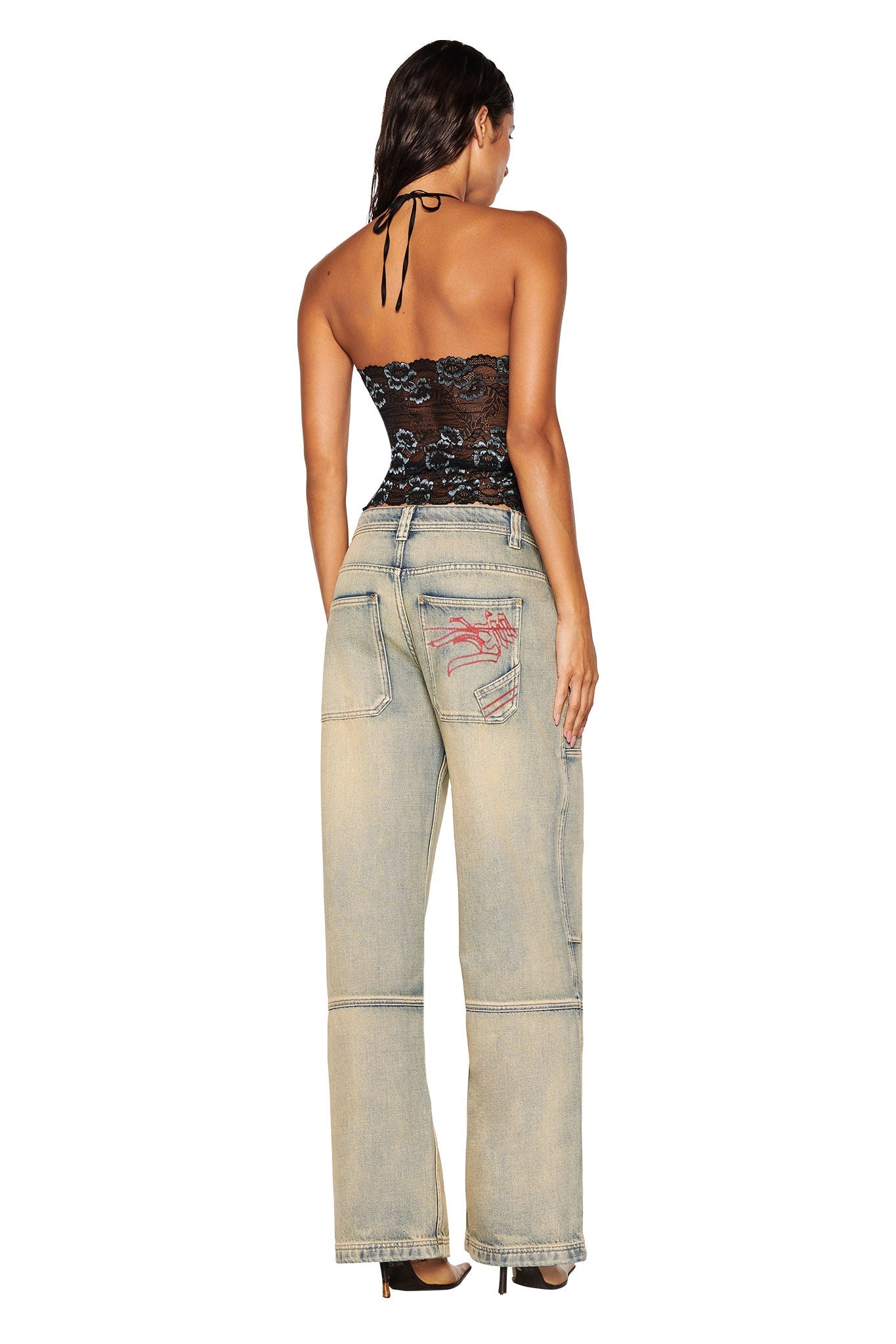 A woman with long dark hair and black heels faces away, wearing a black lace halter top and I.AM.GIA MISTY JEAN - TAN 2.0: loose, light-wash, low-rise jeans featuring patches and red-stitched back pockets.