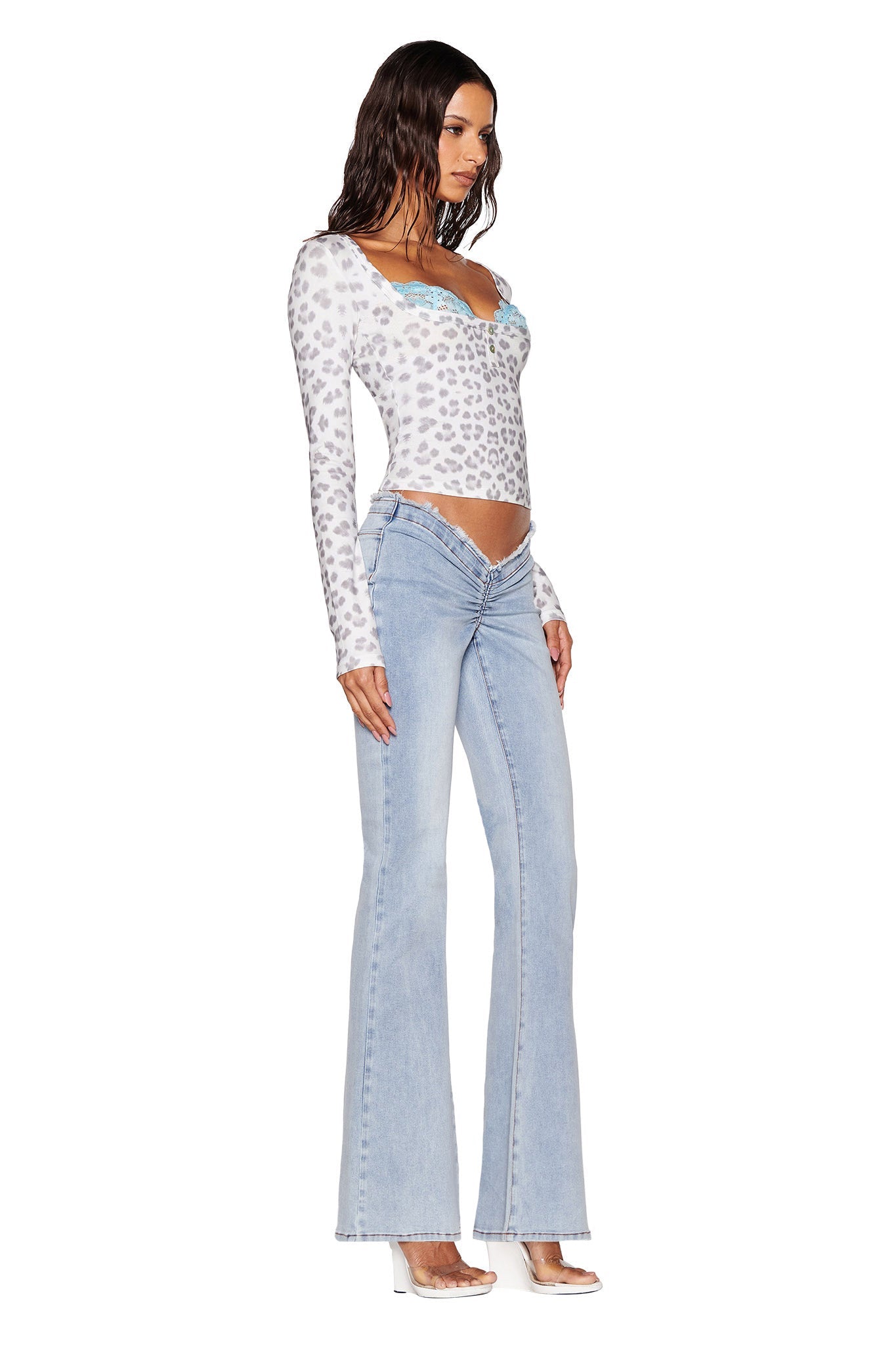 A woman elegantly poses against a white background, wearing the PIPPA TOP - SNOW LEOPARD from I.AM.GIA's Winter Wild Card collection, paired with light blue flared jeans and clear heels.