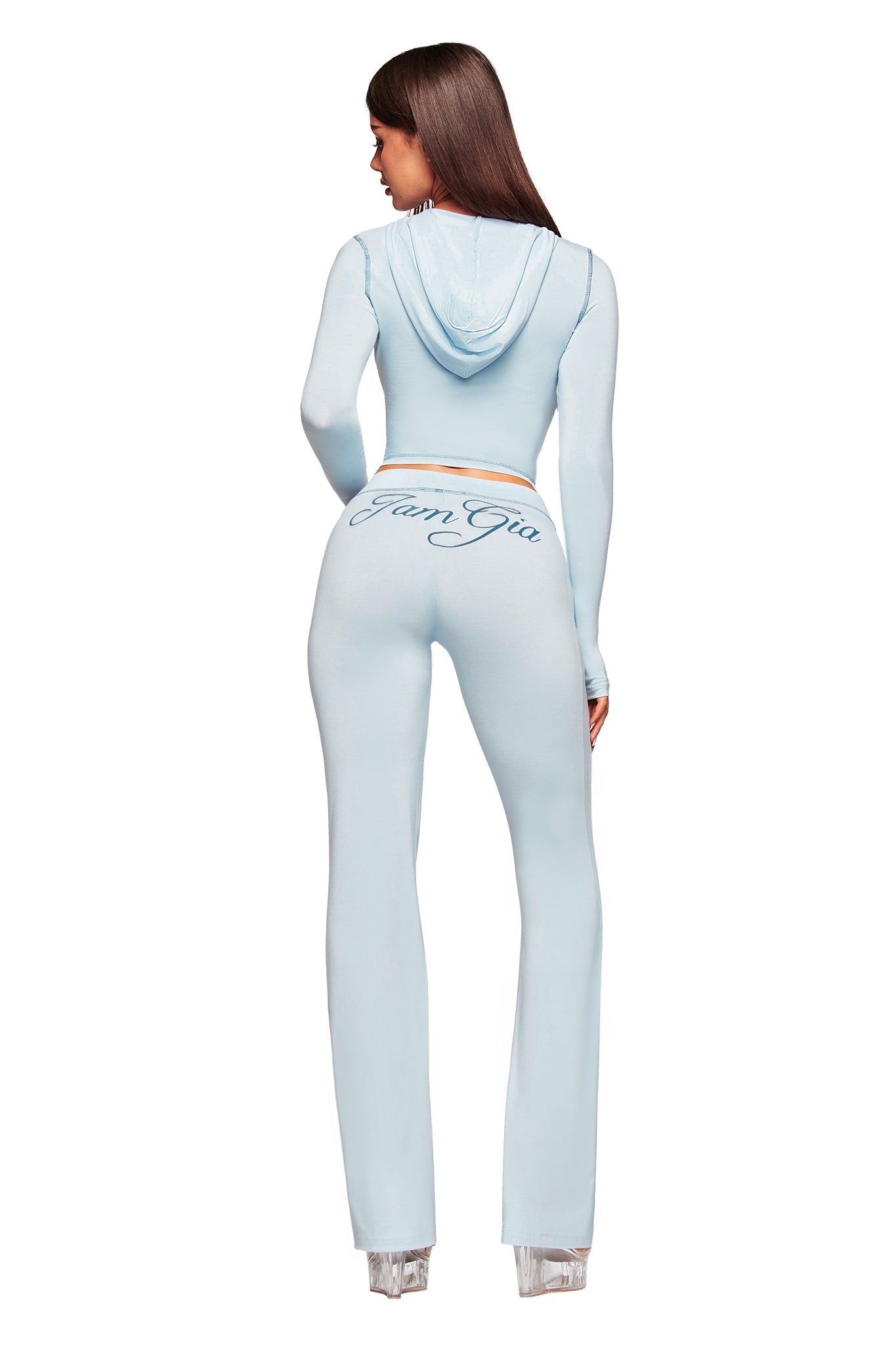A woman faces away in the BLARE HOODIE - BLUE by I.AM.GIA, a baby blue bodycon hoodie tracksuit with a hood. “IamGia” is printed in cursive on the back of her pants. She wears clear high heels, set against a white background.