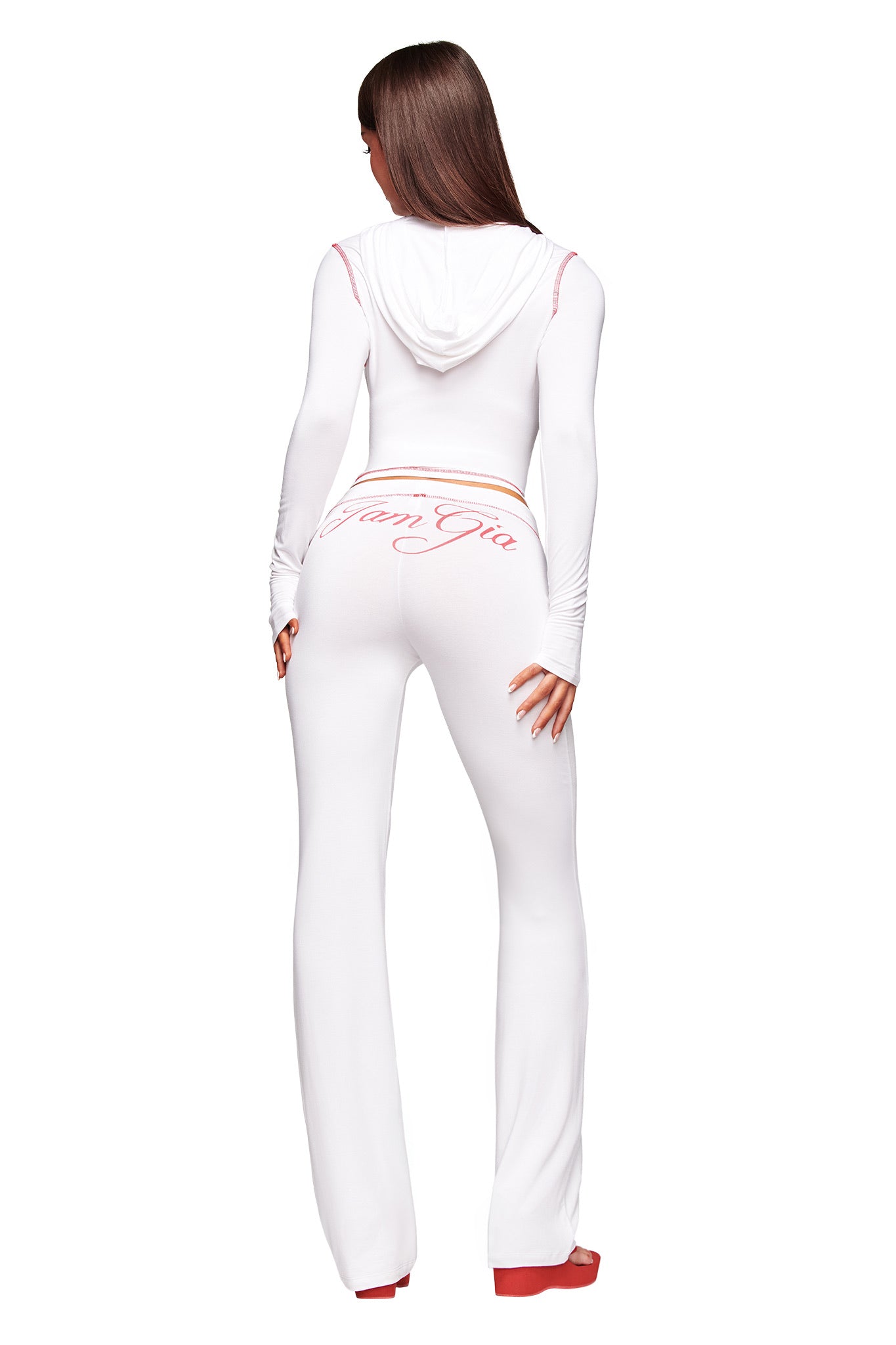 A woman with long straight brown hair faces away, wearing I.AM.GIA's fitted white BLARE TRACKPANT with a hood. "Von Gia" in pink script is across the back. She pairs the look with red platform shoes.