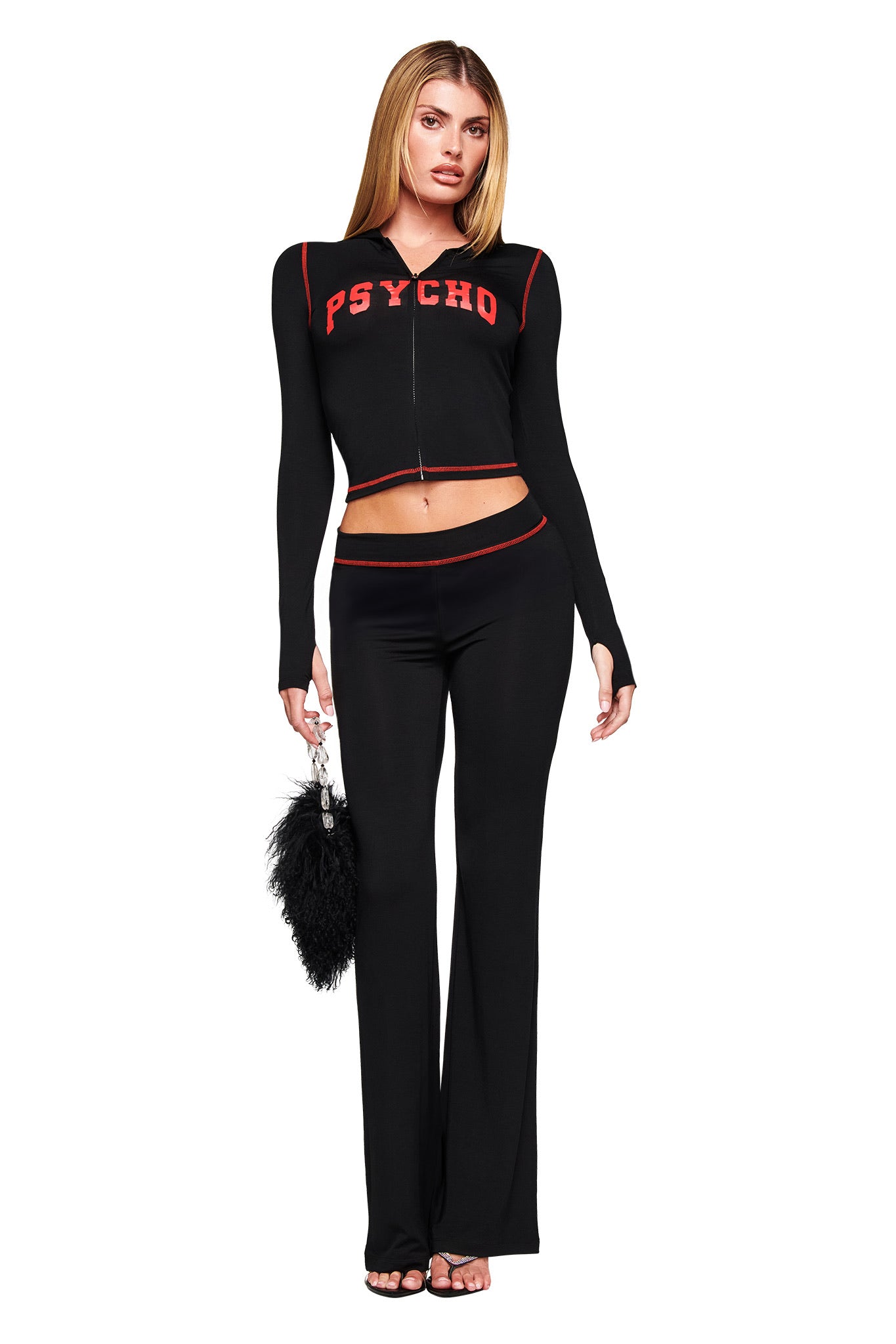 A woman stands against a white background wearing I.AM.GIA's BLARE TRACKPANT - BLACK PSYCHO with the red PSYCHO logo, paired with a matching black crop top and holding a black furry handbag.