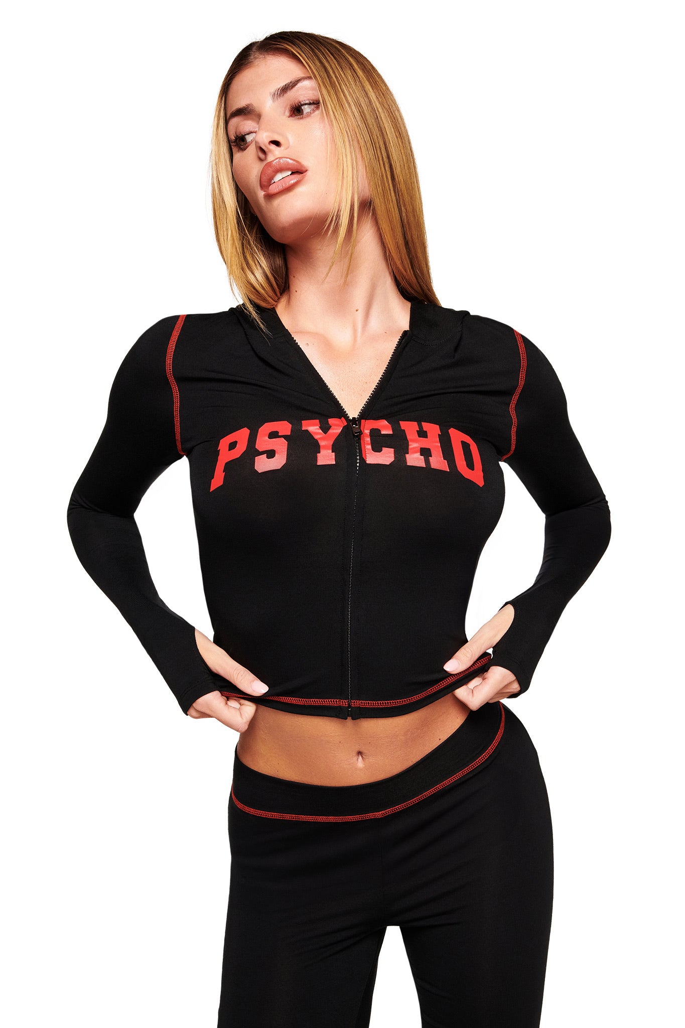 A woman with straight blonde hair wears the I.AM.GIA BLARE HOODIE - BLACK PSYCHO, a black zip-up featuring bold red "PSYCHO" lettering and red piping, paired with matching black pants. She poses tugging at the hoodie’s hem.