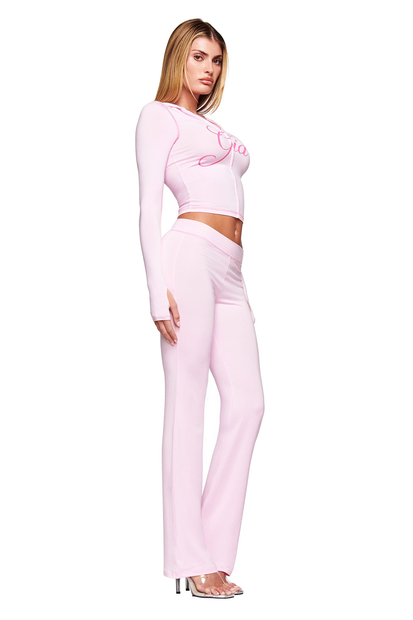 A woman stands sideways against a plain white background wearing the I.AM.GIA BLARE HOODIE in pastel pink and matching high-waisted flared pants with heels.