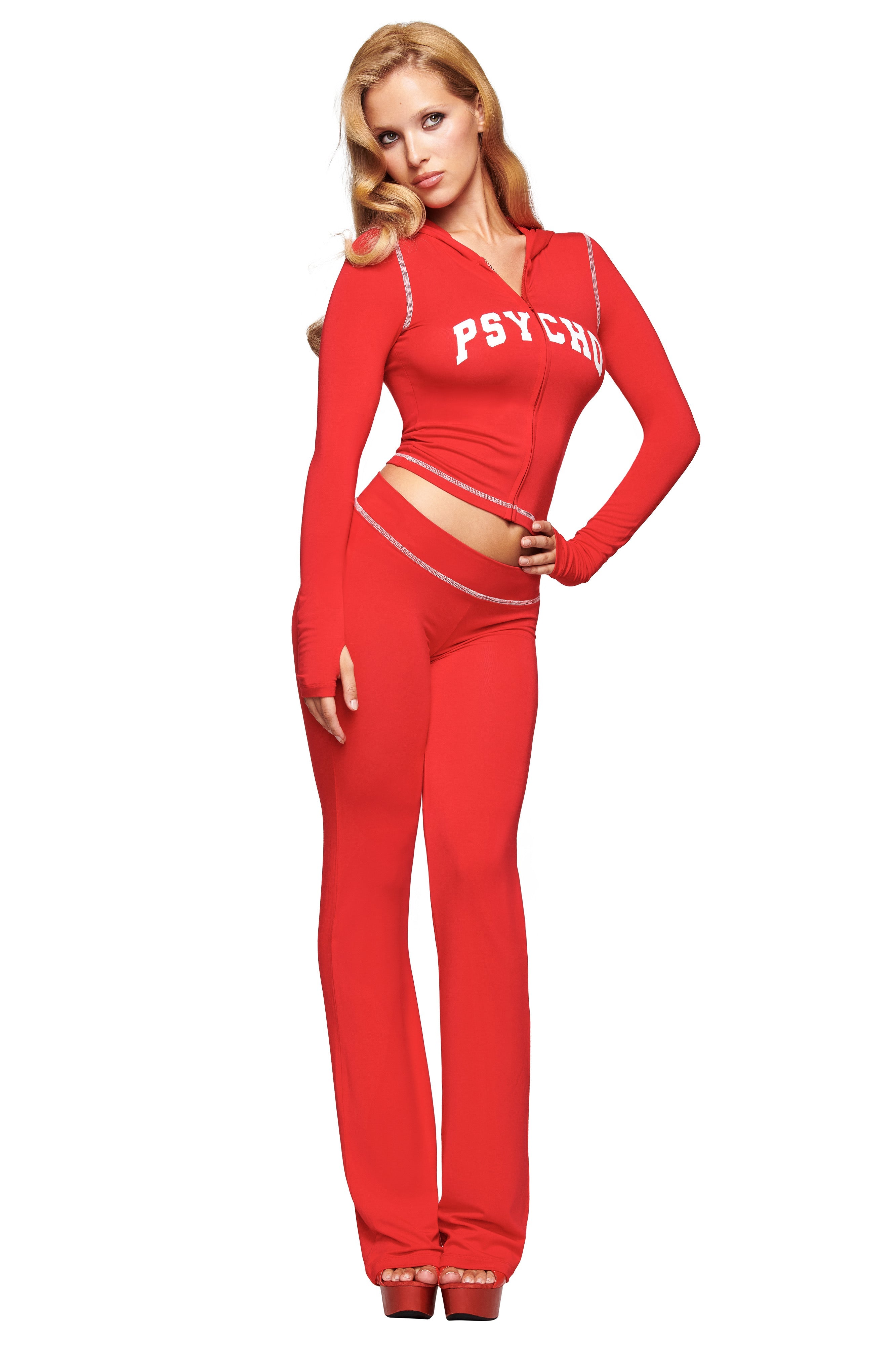 A woman poses in the I.AM.GIA BLARE TRACKPANT - RED : PSYCHO : SALSA, featuring low-rise, flared legs, cutout waist details, long sleeves, and "PSYCHO" on the chest; she has long blonde hair and wears high heels.