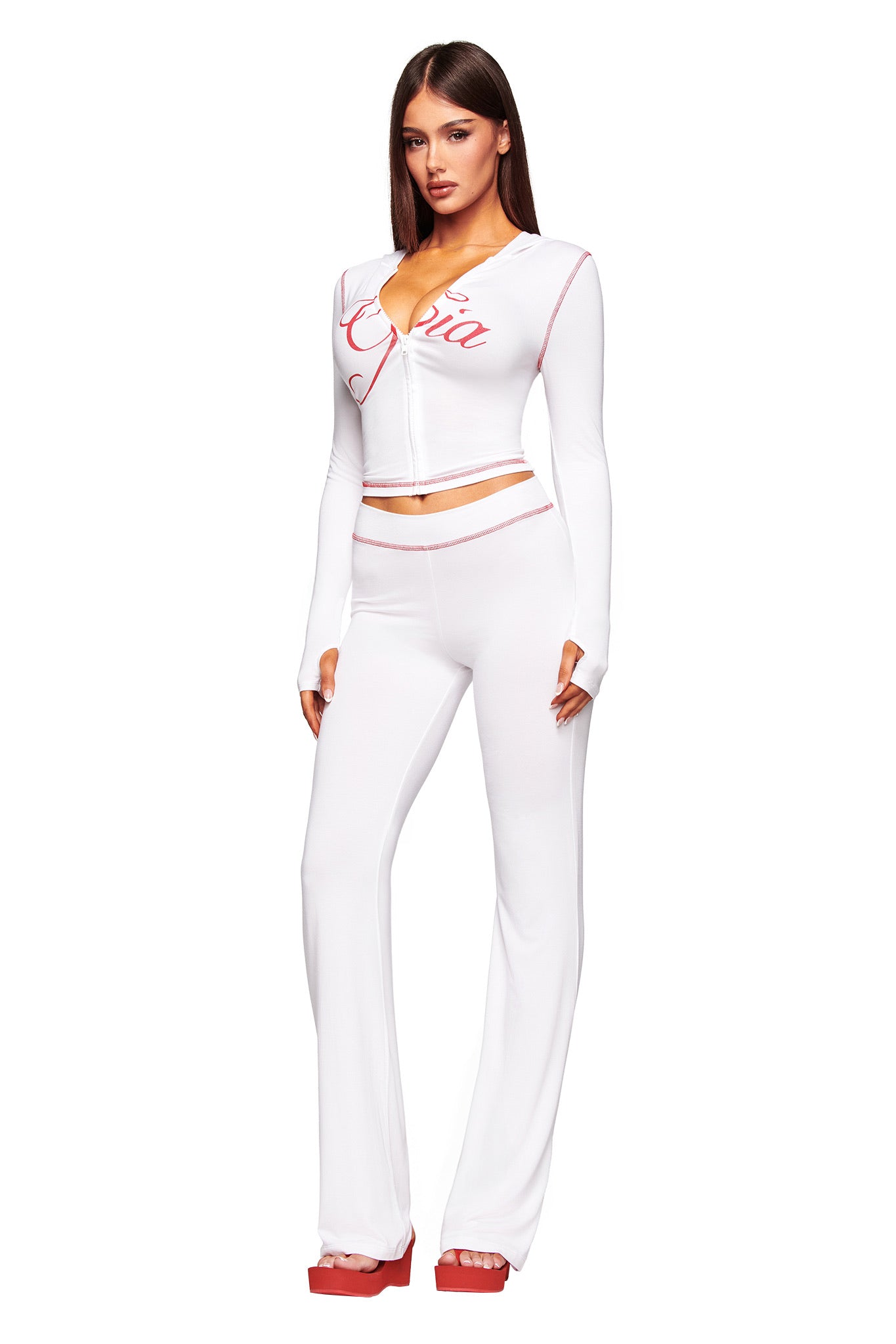 A woman wears a fitted long-sleeve white crop top with red trim and lettering, paired with I.AM.GIA's BLARE TRACKPANT - WHITE high-waisted flared pants and red platform heels, against a plain white background.