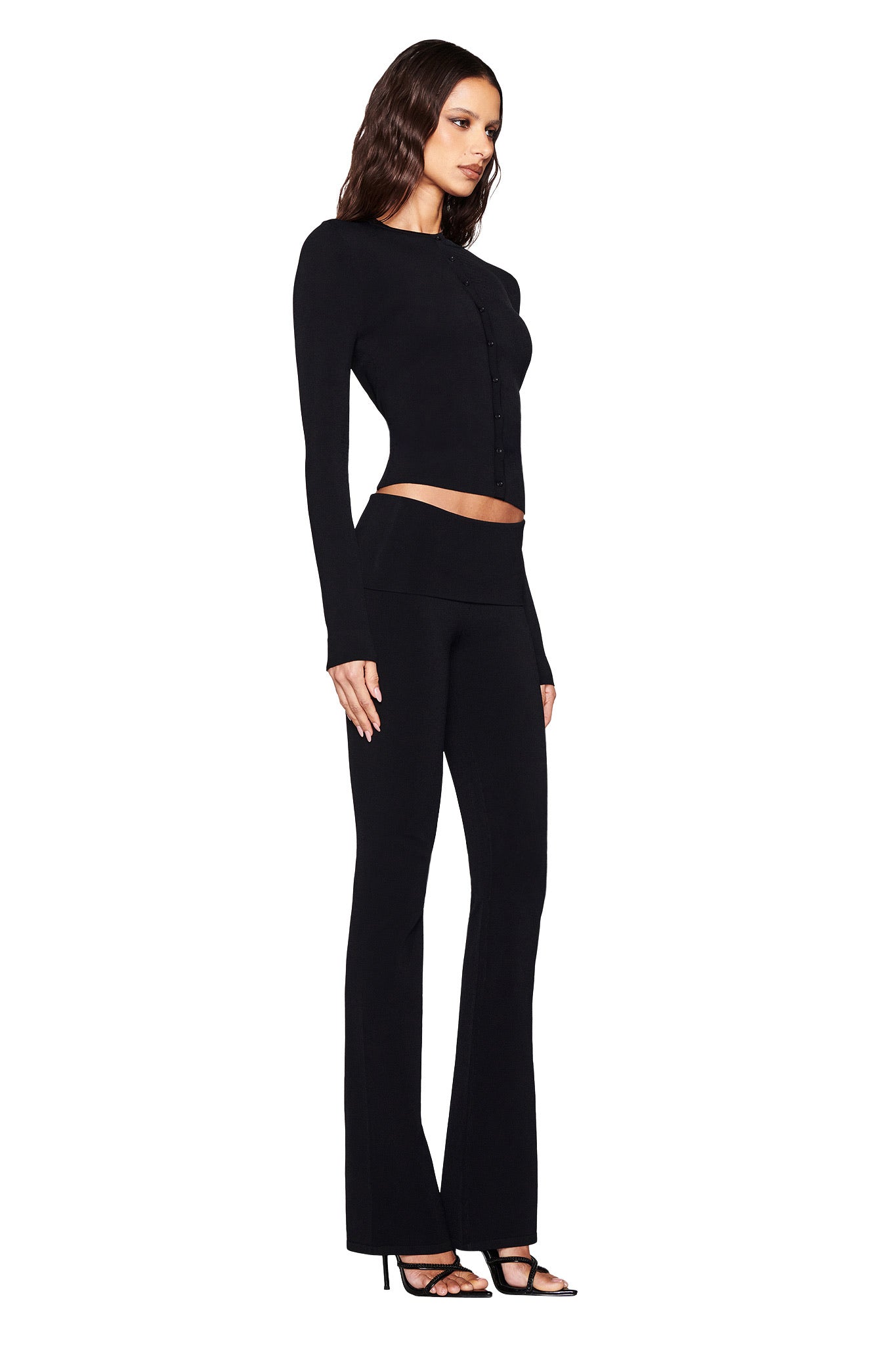 A person stands in profile wearing the I.AM.GIA AUDRINA CARDIGAN 2.0 – BLACK and black flared pants, highlighting their wavy hair and high-heeled sandals against a white background.