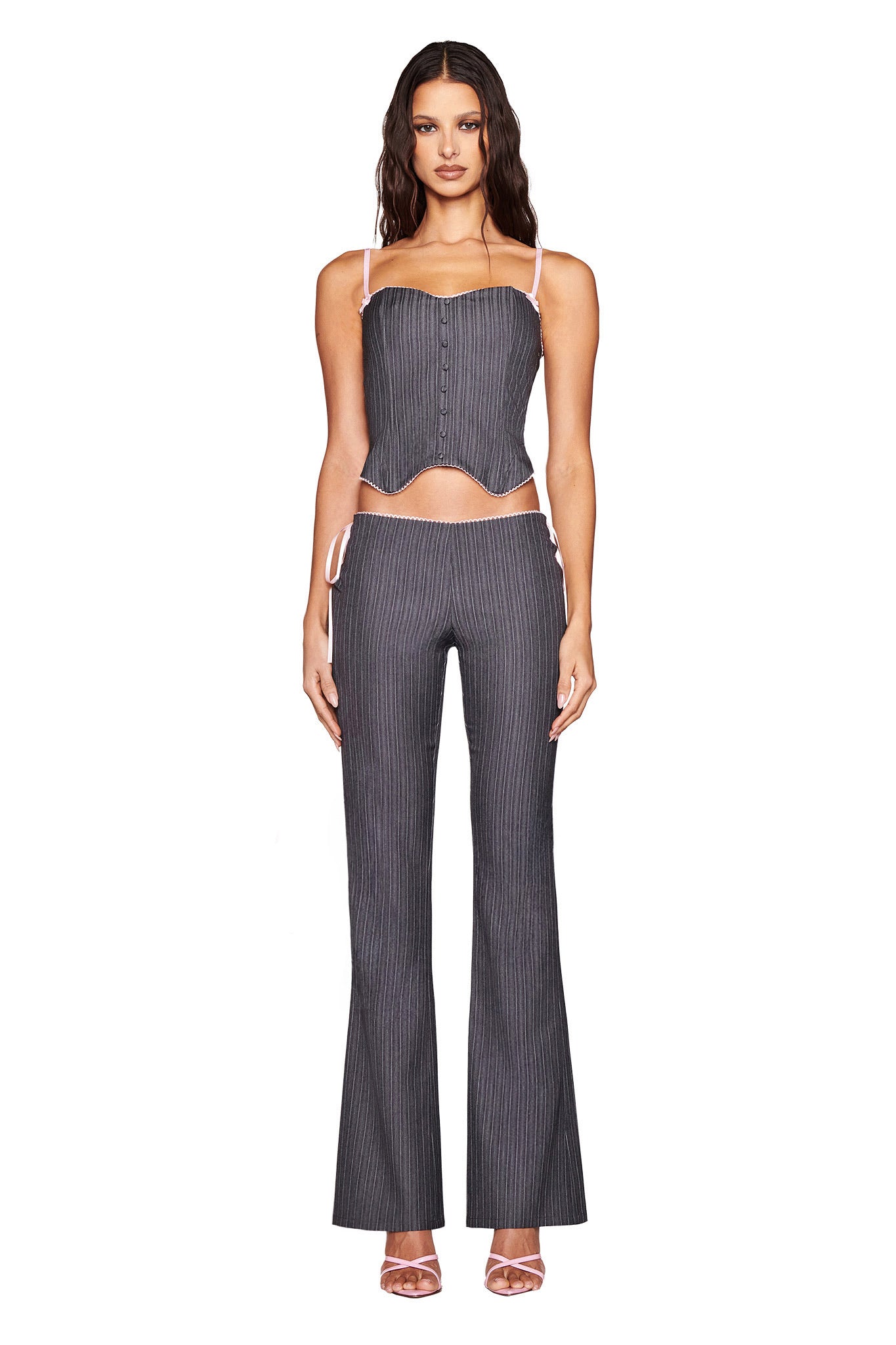 A woman models the I.AM.GIA FLORIAN CORSET in grey pinstripe with adjustable straps, paired with matching pants and pink open-toe heels. The chic outfit is accentuated by front buttons and set against a white background.