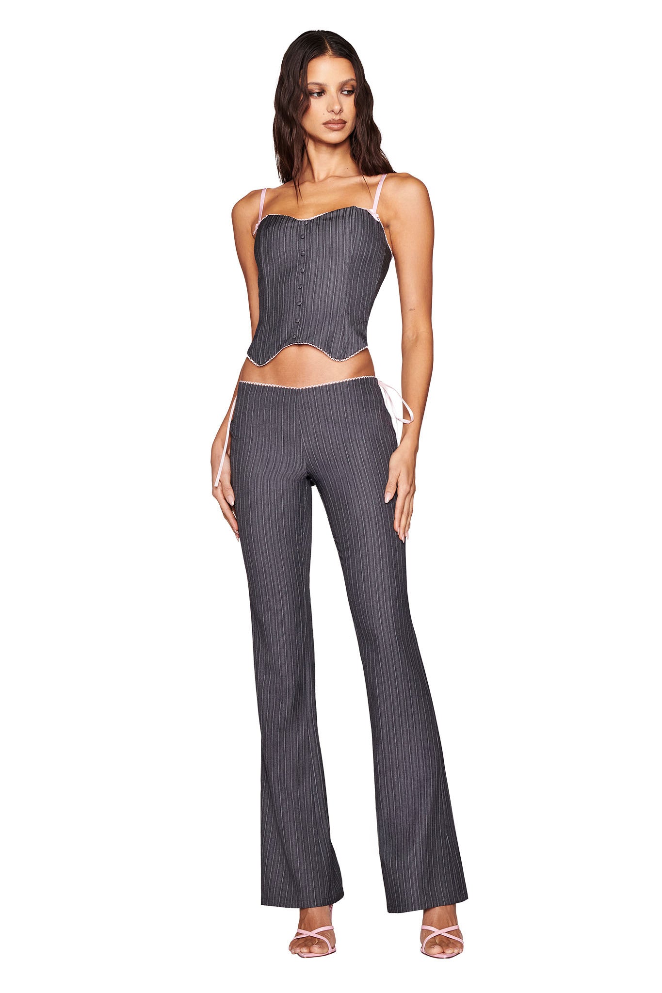 A person wears the I.AM.GIA FLORIAN PANT - GREY pinstripe set with a matching corset top featuring thin straps and side lace-up details, paired with open-toed heels against a plain white background.