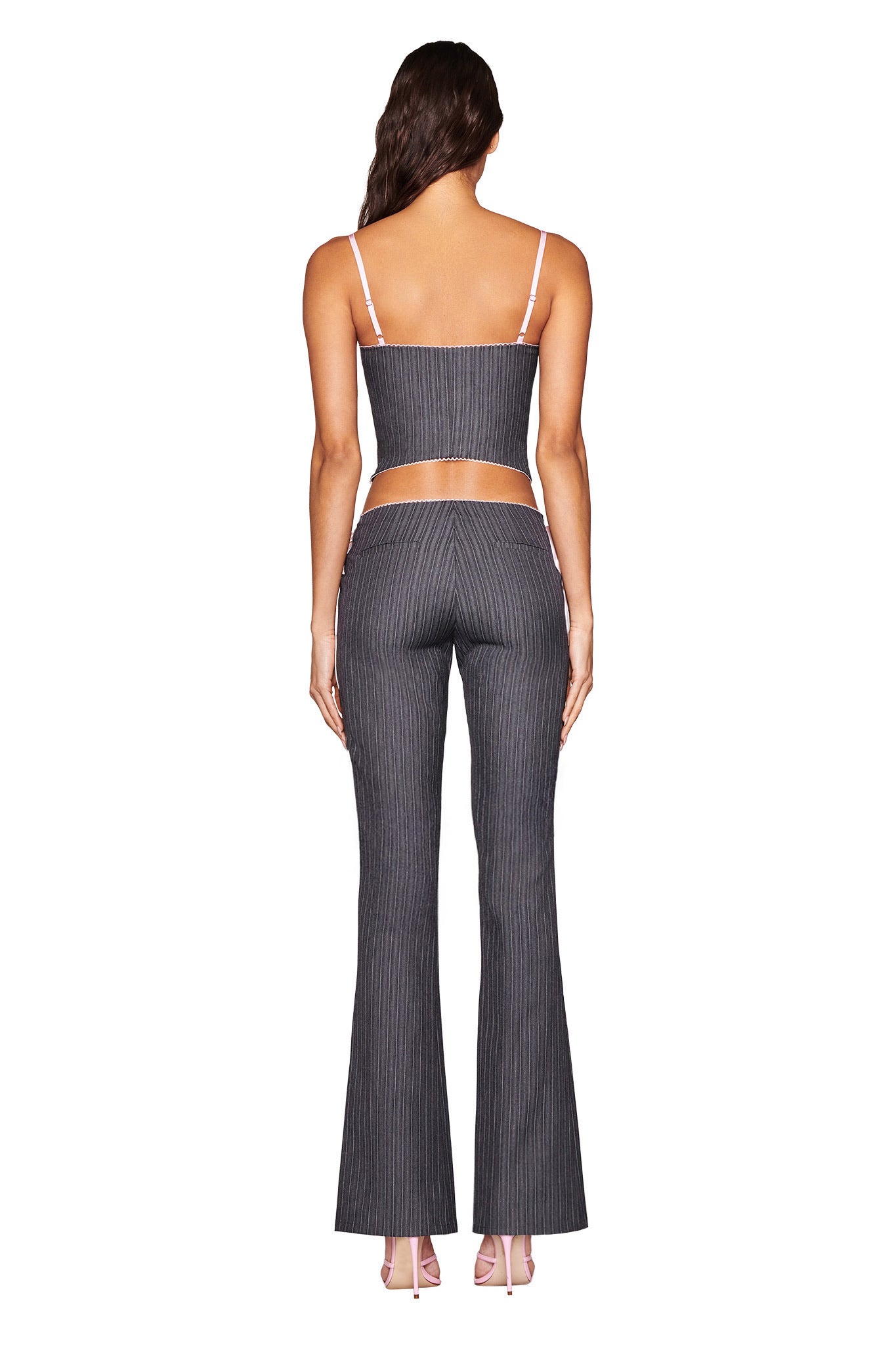 A person is facing away on a white background, wearing the FLORIAN PANT - GREY pinstripe outfit by I.AM.GIA, featuring flared leg pants and a sleeveless top with pink lace-up details, paired with light pink high heels.