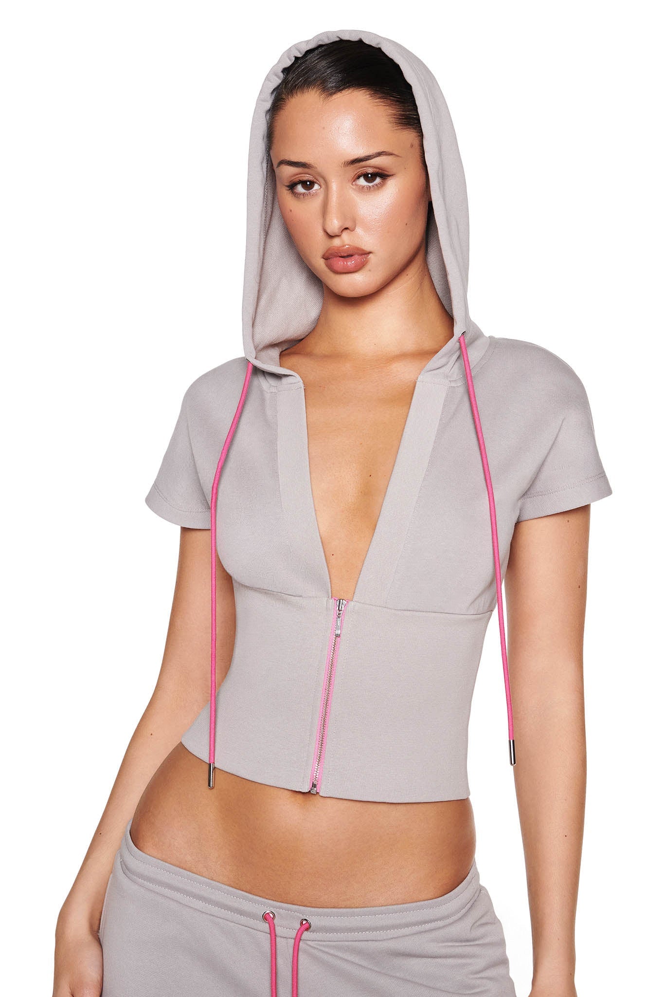 A person models the ARLO TRACK JACKET - GREY from I.AM.GIA, a chic zip-up with short sleeves, a hood, and deep V-neck accented by neon pink drawstrings, paired with matching pants made of soft French terry fabric against a plain white background.