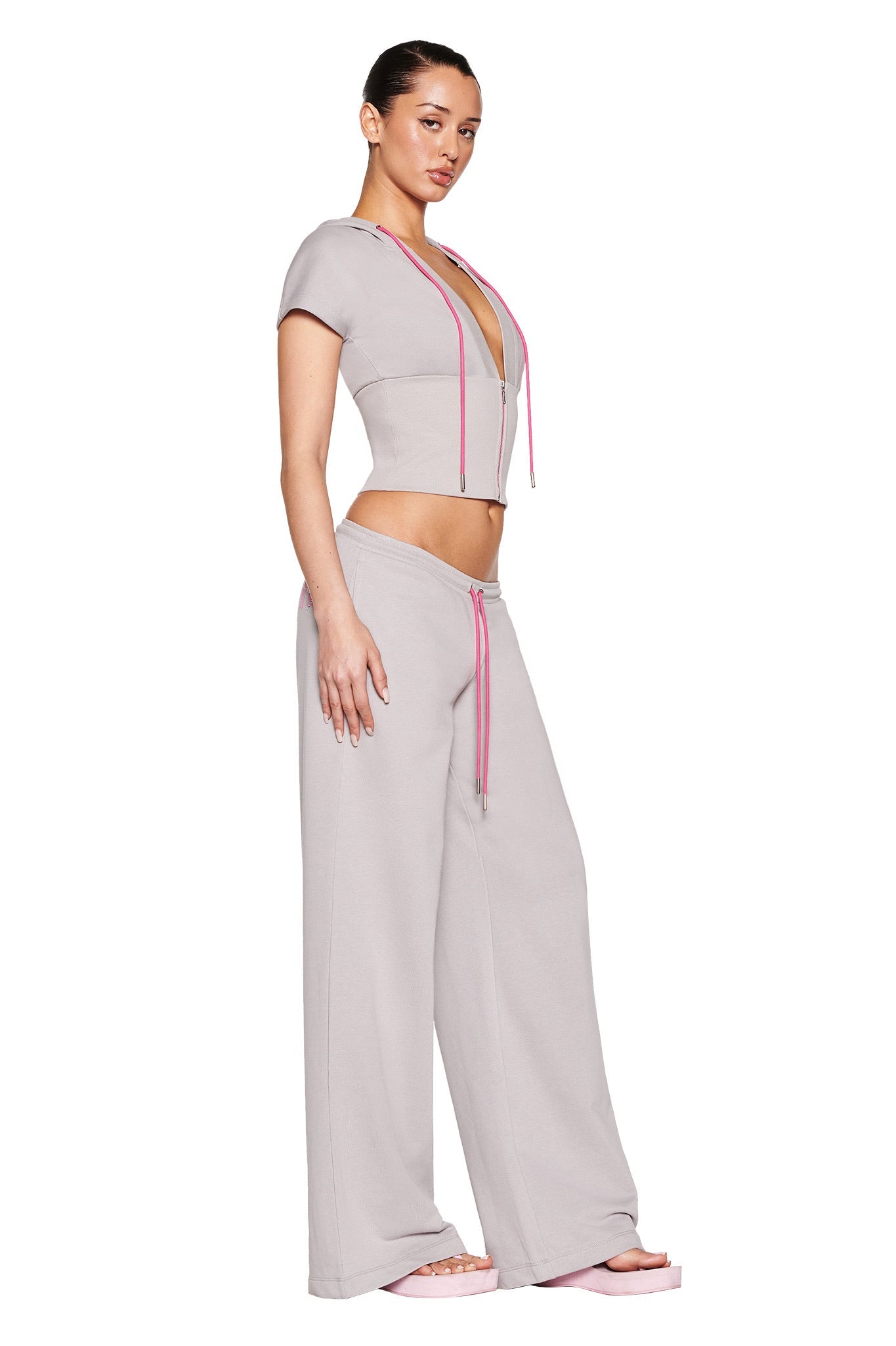 A person confidently poses in an I.AM.GIA ARLO TRACK PANT - GREY : PINK, featuring a fitted gray crop top with pink drawstrings and high-waisted, wide-leg track pants. They're wearing pink platform sandals with slicked-back hair against a white background.