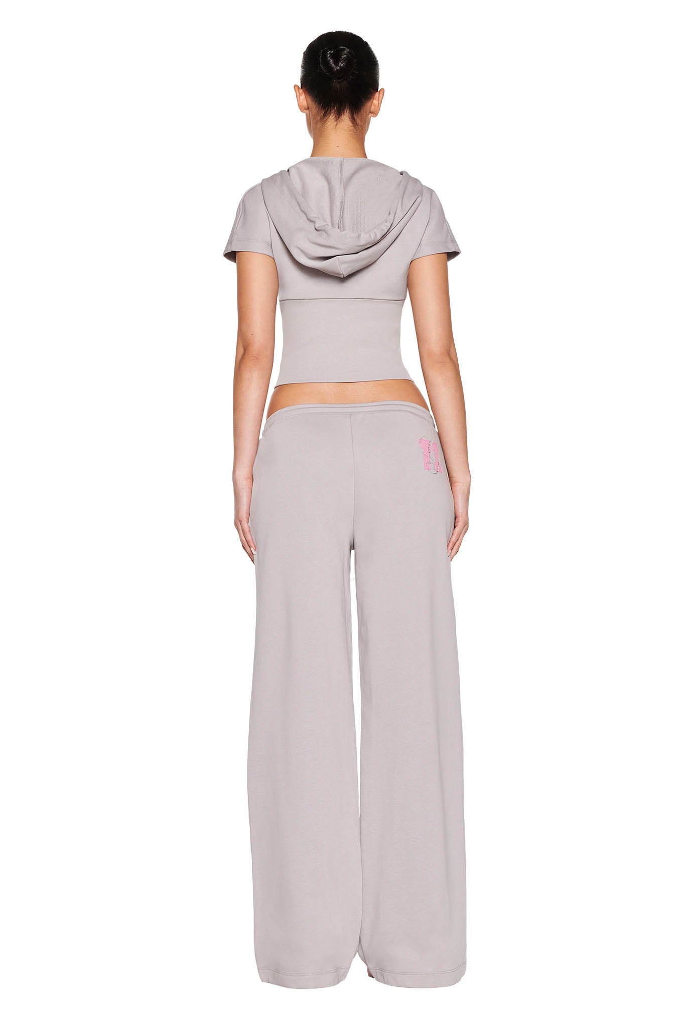 Person in a light gray ensemble stands with back turned, wearing the ARLO TRACK JACKET - GREY by I.AM.GIA. The cropped short-sleeve hoodie features a neon pink drawstring. Hair is styled in a bun against the plain white background.