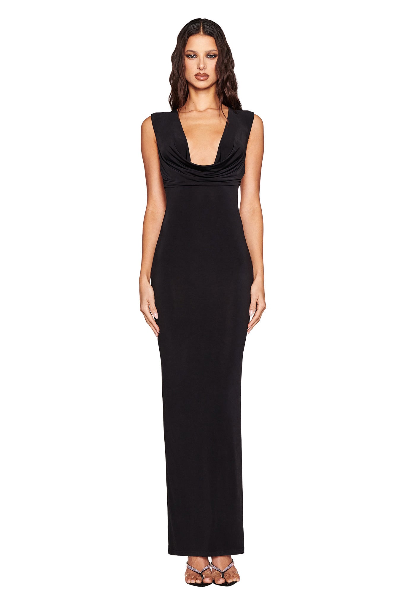 A woman poses against a white background in the I.AM.GIA MILANI MAXI DRESS, featuring a plunging cowl neckline and cap sleeves. Her long, dark wavy hair and strappy heels complete her timeless, elegant look.